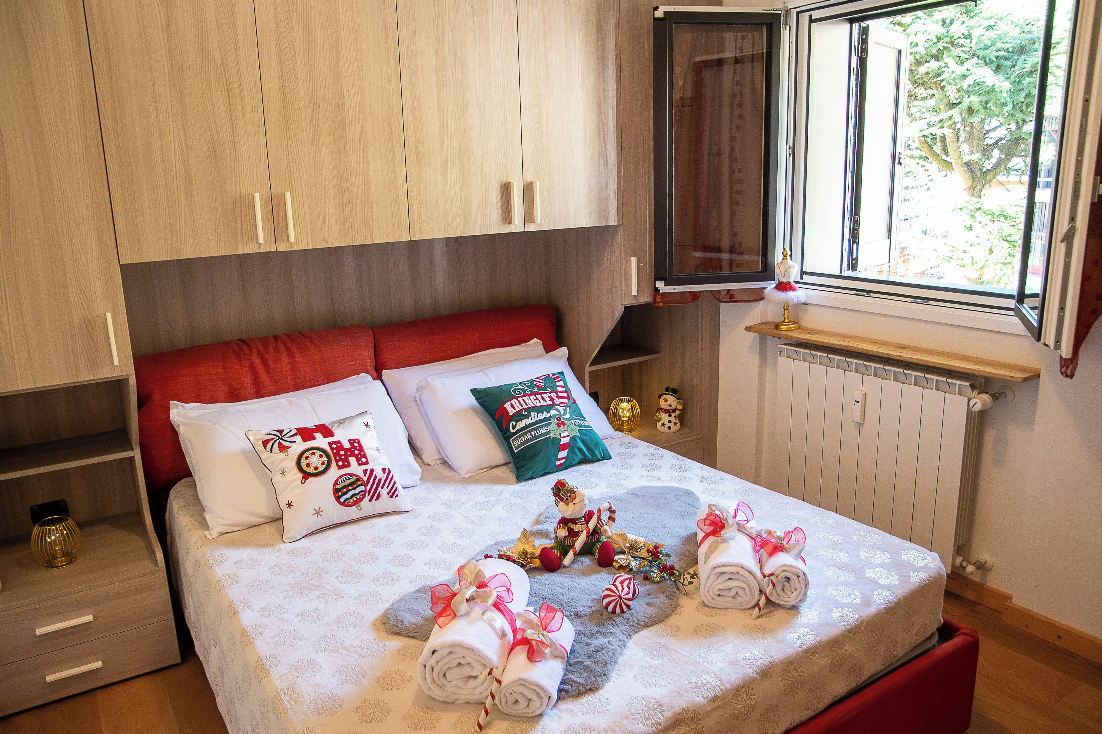 Een net opgemaakt bed met een rode hoofdbord, witte beddengoed en decoratieve kussens. Op het bed liggen opgerolde handdoeken, een kleine kerstboom, en een raam met uitzicht op bomen.