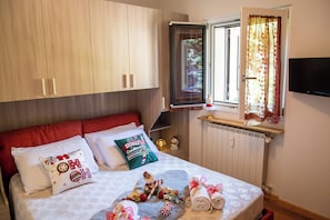 Ein ordentlich bezogenes Bett mit rotem Kopfteil, ein Fenster mit Vorhängen und ein Fernseher an der Wand.