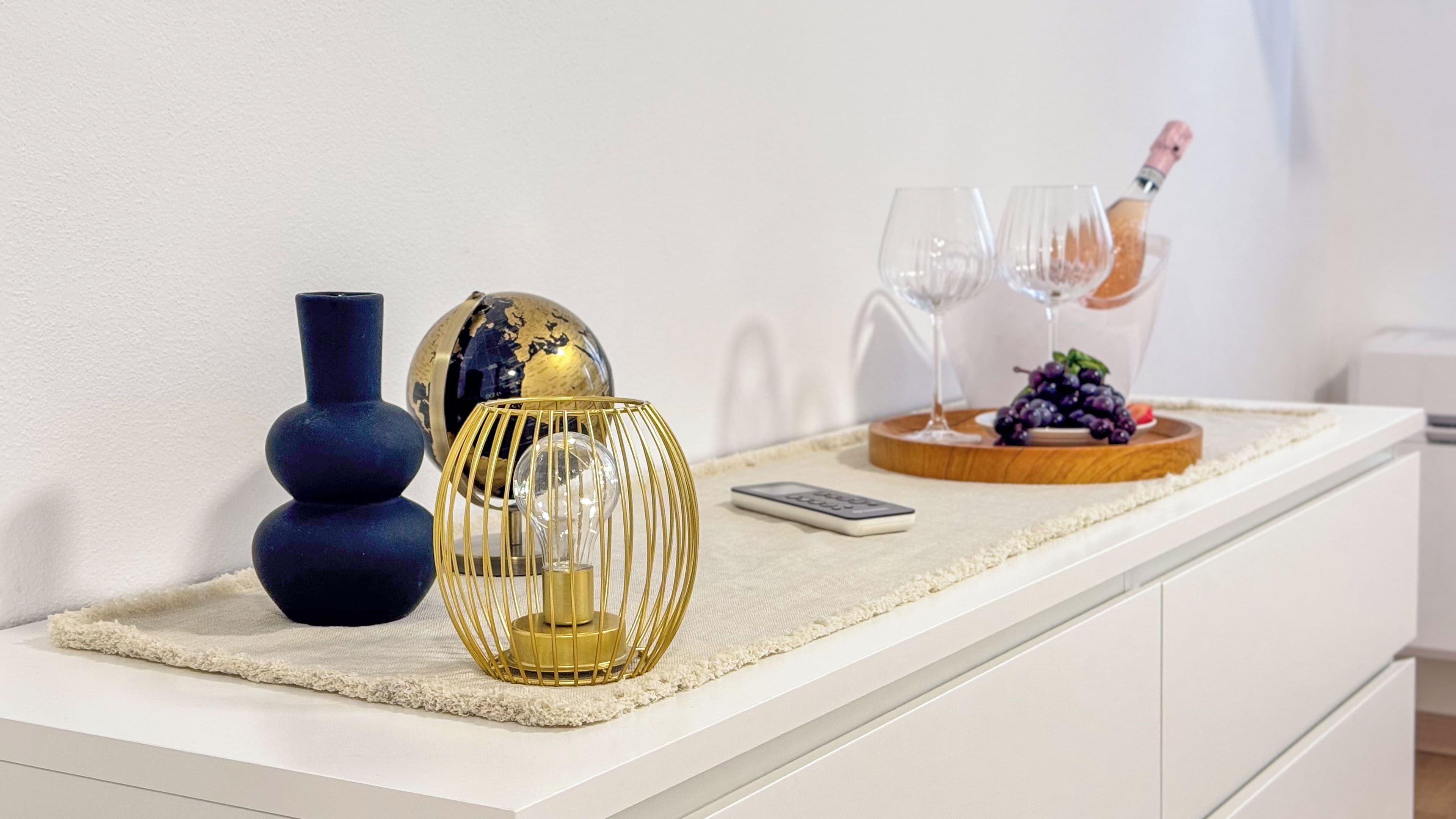 A white countertop with a globe, a vase, a candle holder, a remote control, and a tray with a glass and grapes.