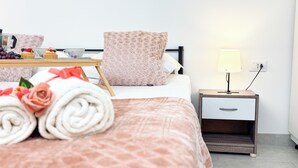 Una cama bien hecha con toallas enrolladas y una almohada decorativa, una mesita de noche con una lámpara y un frutero pequeño con uvas y arándanos.