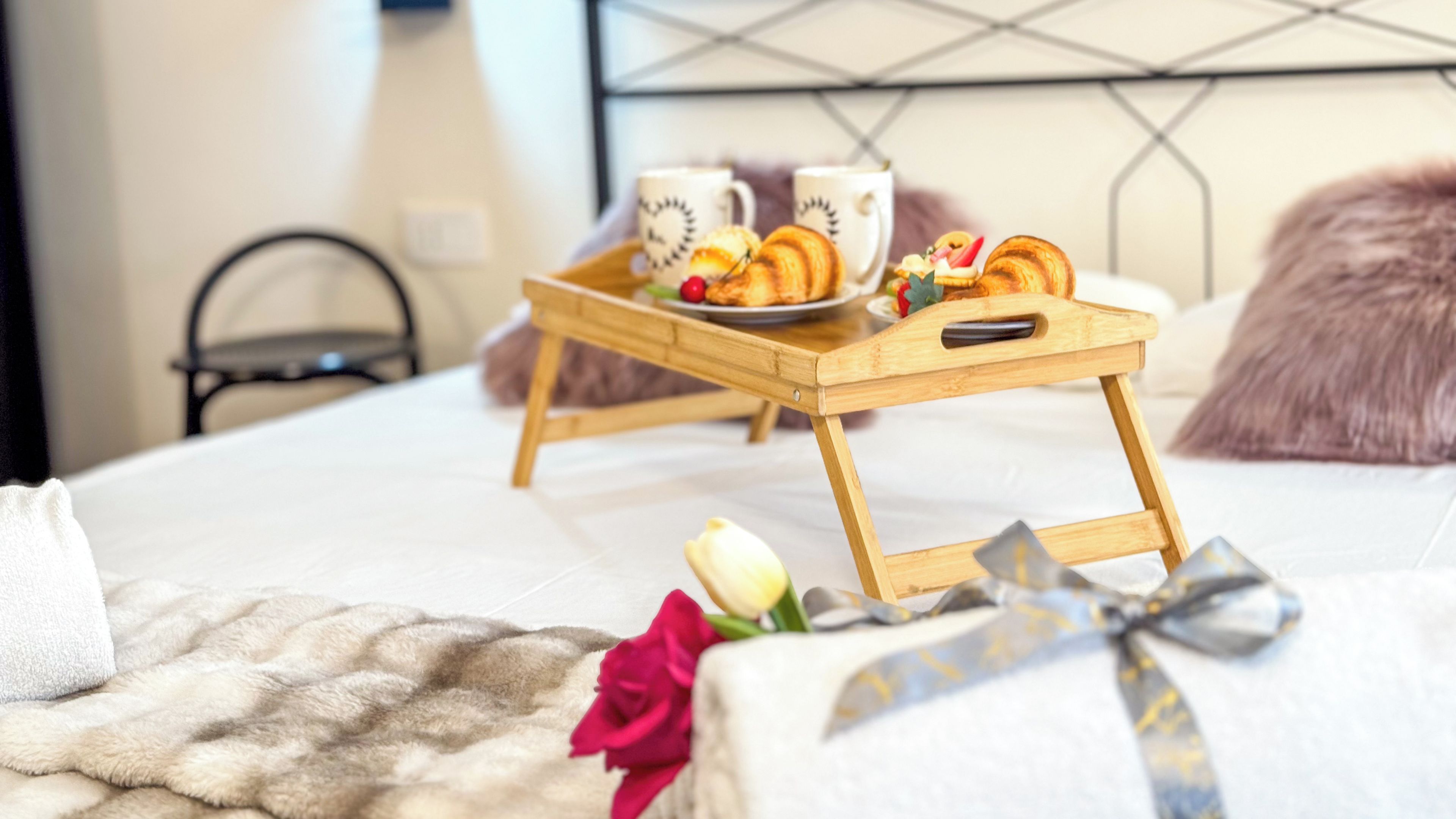 A neatly made bed with a wooden tray holding breakfast items, a bouquet of flowers, and a gift on a fluffy white blanket.