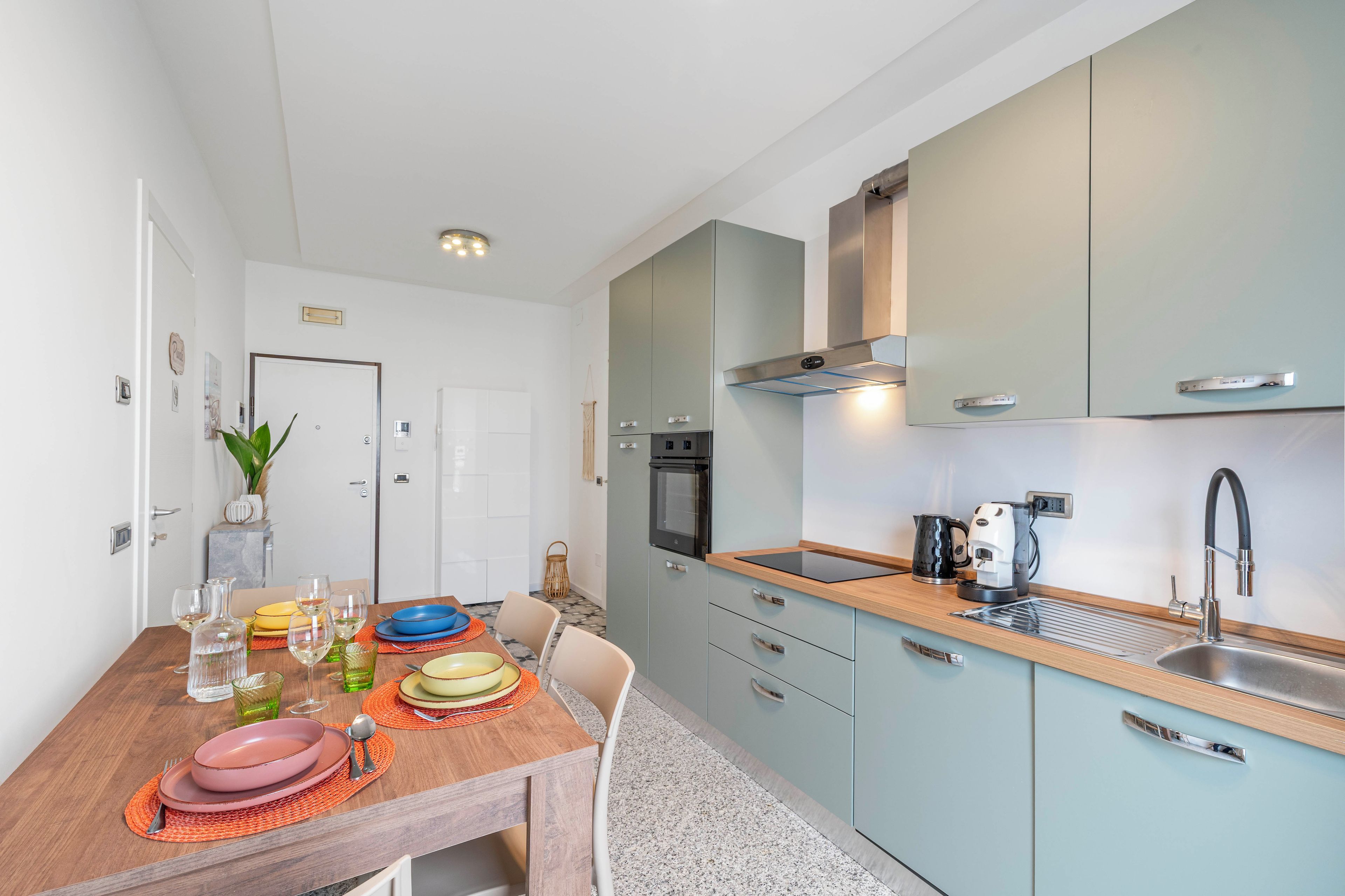 Una cocina moderna con una mesa de comedor de madera dispuesta para cuatro personas, un horno empotrado y un lavamanos de acero inoxidable.