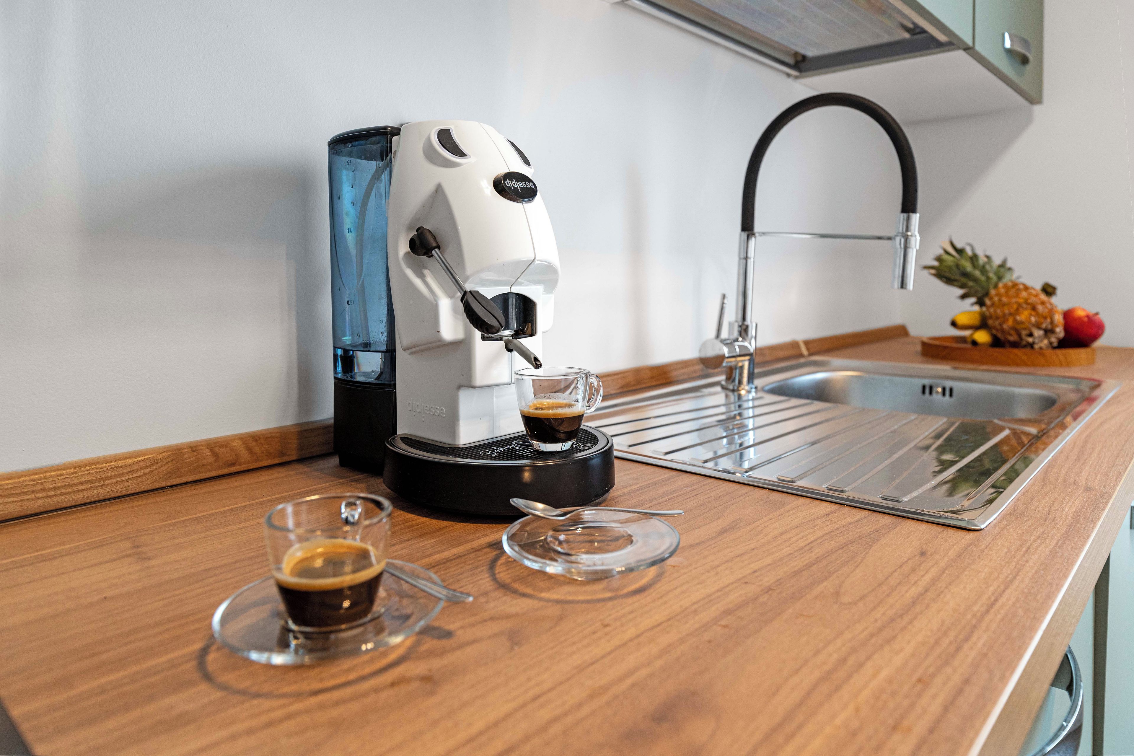 Una cafetera sobre una encimera de madera con dos tazas de café, un vaso y un frutero.