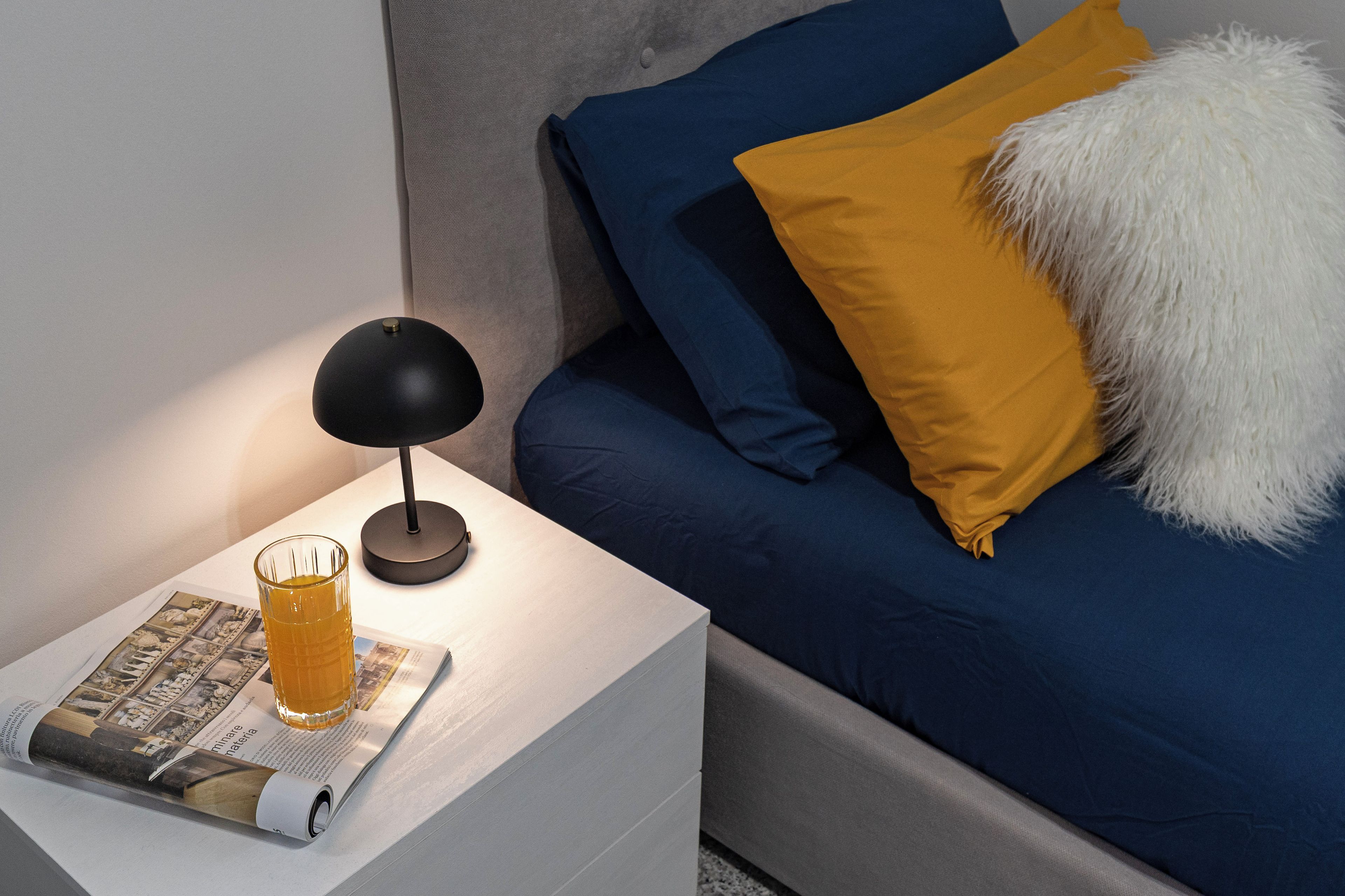 A neatly made bed with blue and yellow pillows, a white fluffy pillow, and a glass of orange juice on a white nightstand beside a magazine.