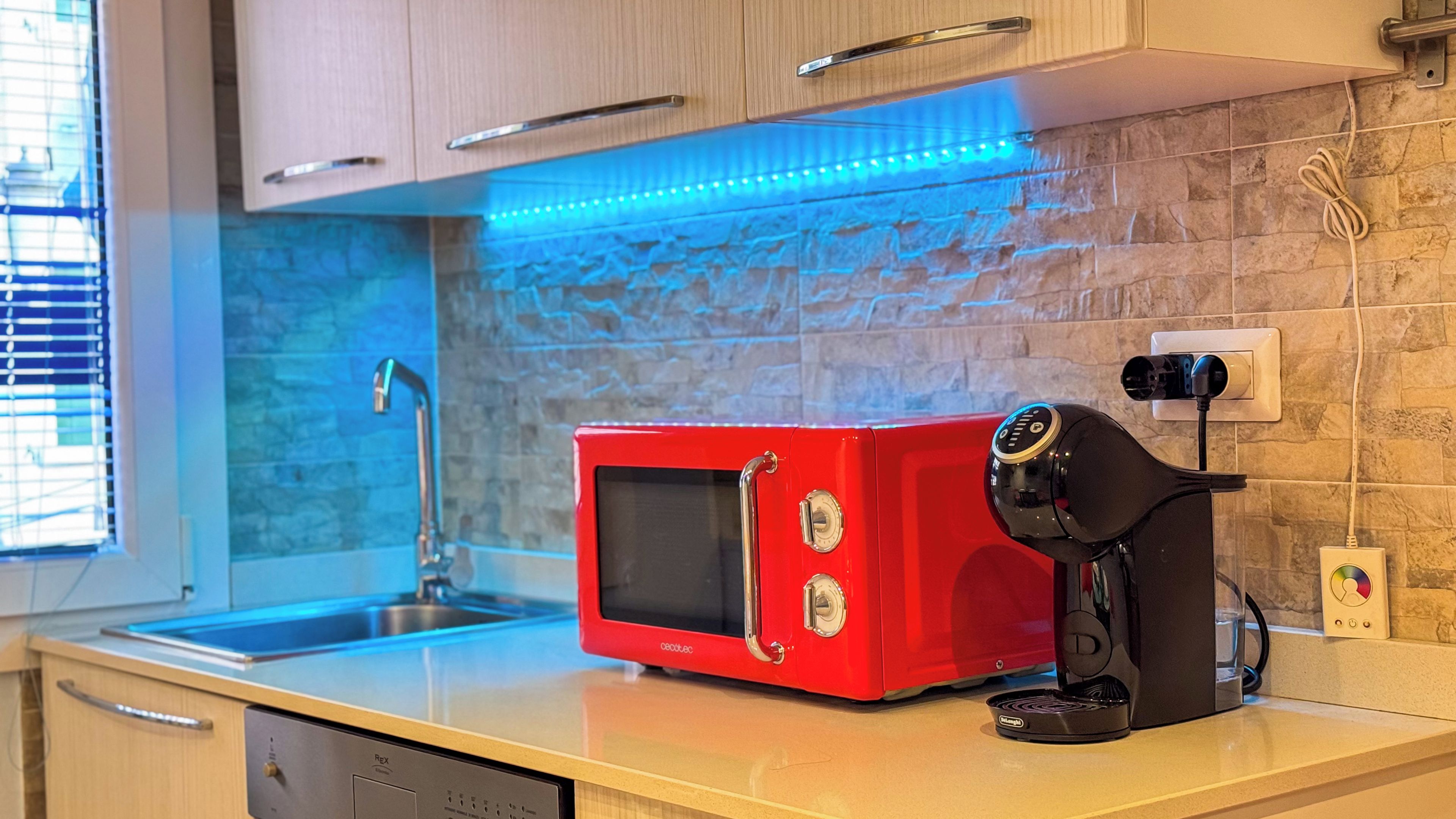 Une cuisine avec un micro-ondes rouge, une machine à café, un évier et une fenêtre avec des stores.