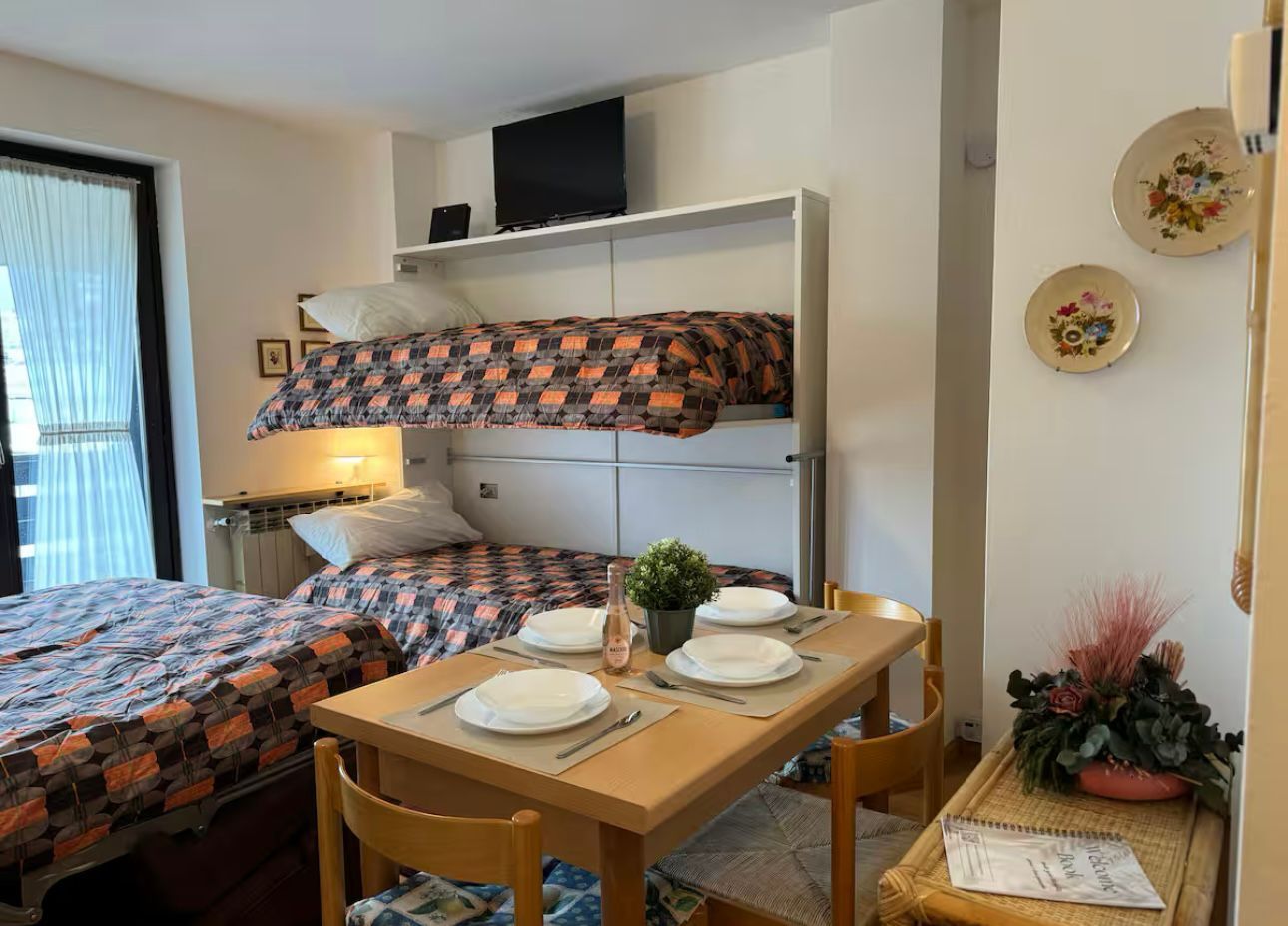 A compact hotel room with a bunk bed, a dining table set for two, and a small side table with a plant.