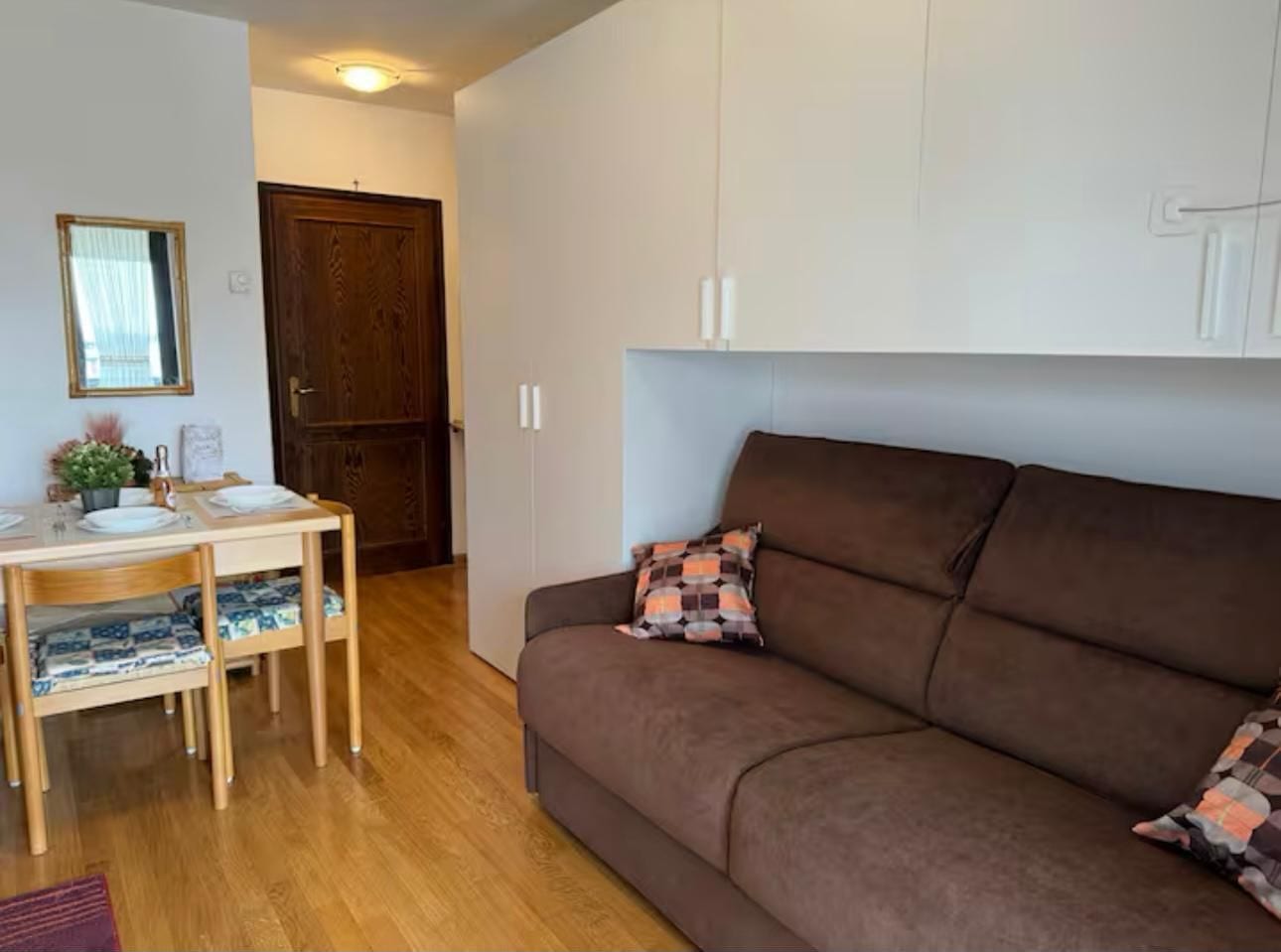 A cozy living room with a brown sofa, a wooden dining table with chairs, and a small table with decorative items.