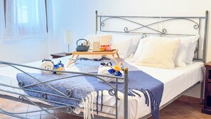 Cama bem arrumada com estrutura de metal, roupa de cama branca e um cobertor listrado em azul e branco. Uma bandeja de madeira com uma chaleira, xícara e um pequeno buquê de flores está colocada sobre a cama.