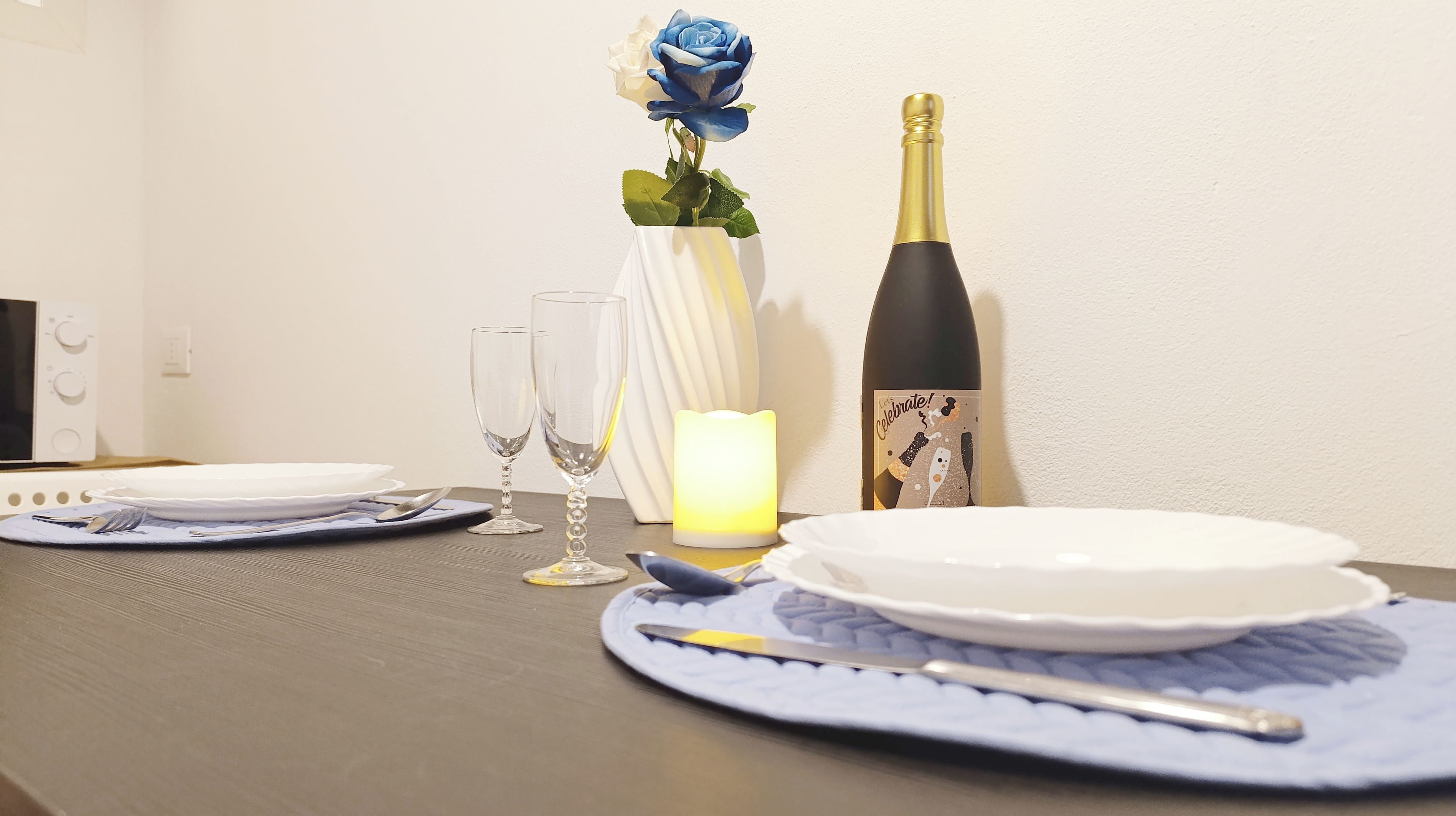 Una mesa de comedor dispuesta con platos, cubiertos, una vela, una botella y un jarrón con una rosa azul.