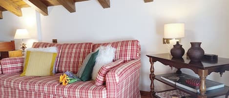 Un sofá rojo con estampado de cuadros y almohadones decorativos, una mesa de madera con un jarrón y una lámpara.