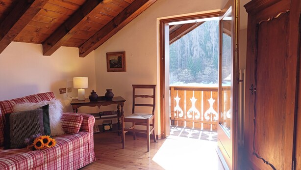 Quarto acolhedor com teto de madeira, sofá xadrez, uma cadeira, uma mesinha com abajur e vista para uma paisagem nevada através de uma porta.