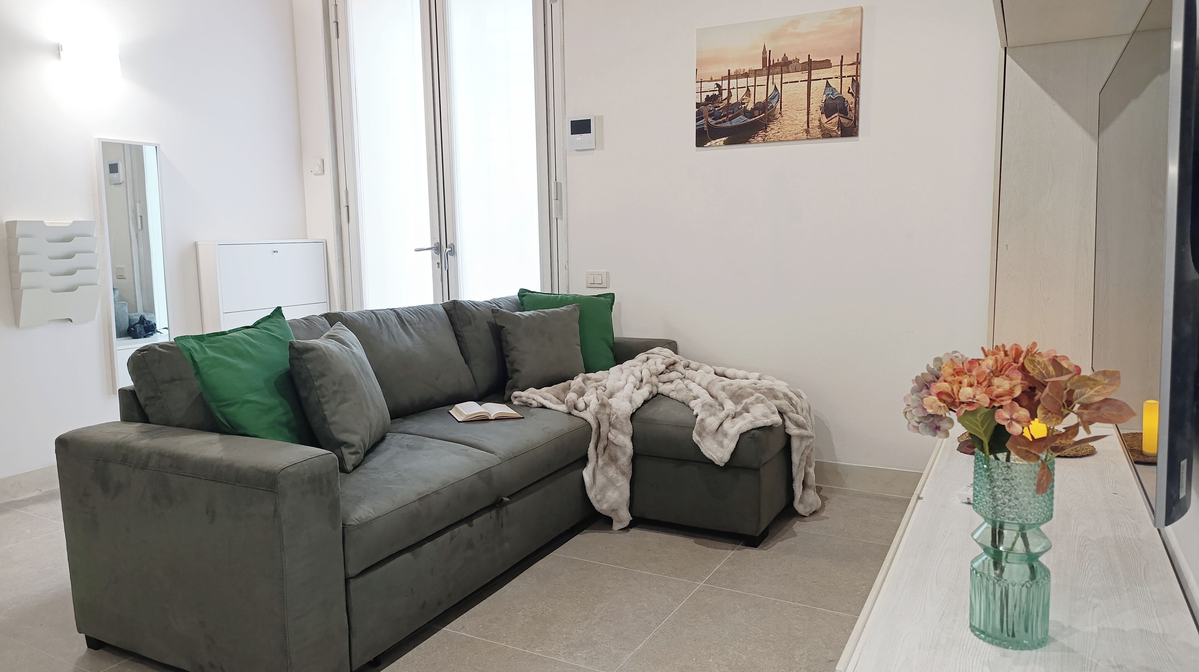 Uma sala de estar moderna com um sofá cinza, almofadas verdes e cinzas, um cobertor e um vaso com flores.