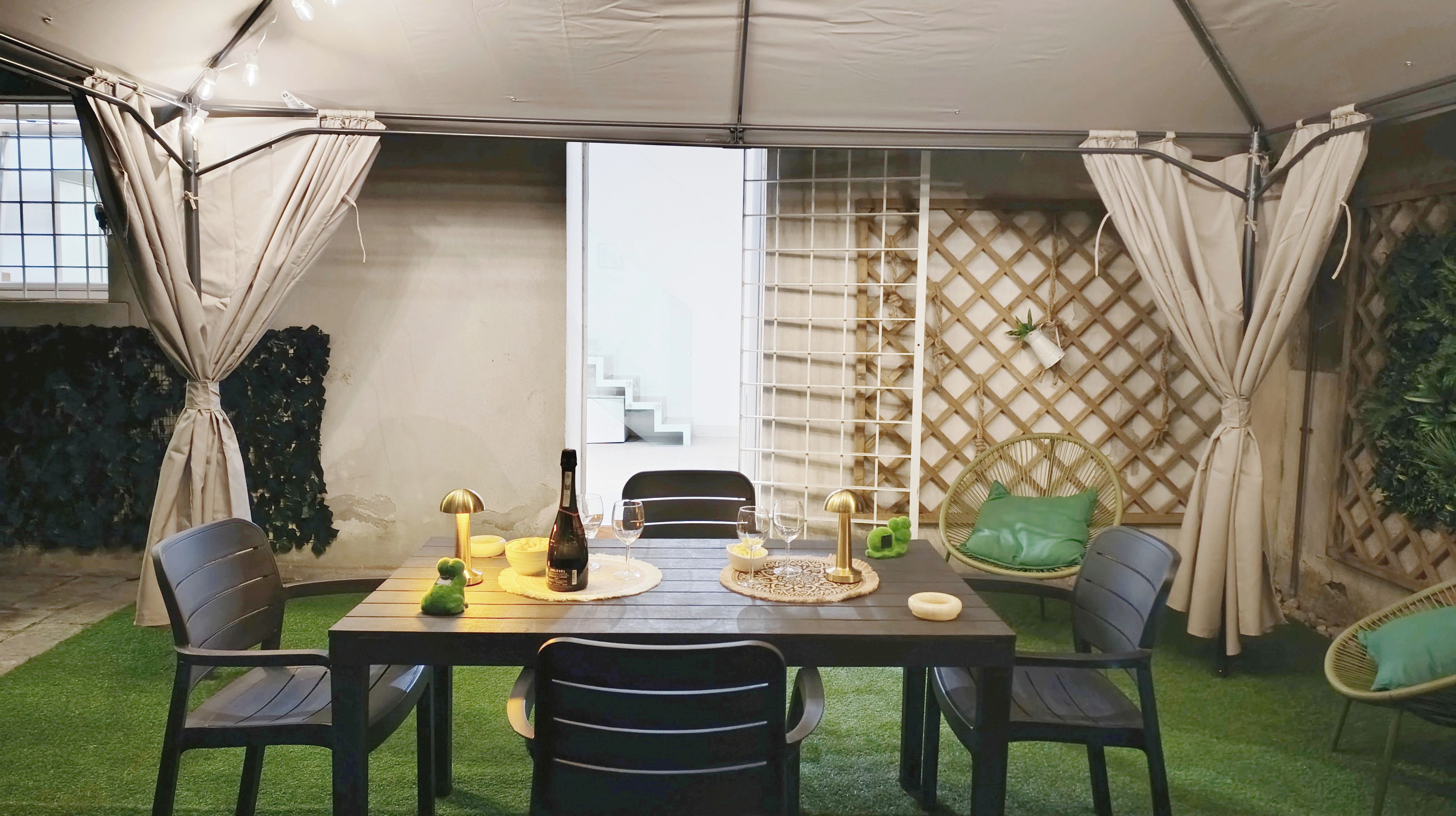A tented outdoor dining area with a table set for four, surrounded by chairs, and a wicker chair with a cushion.