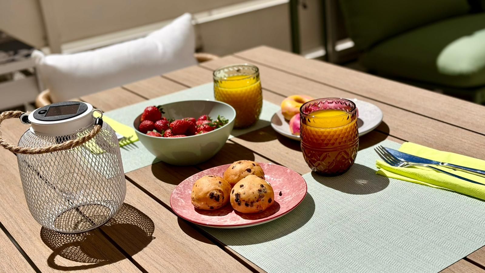 Uma mesa de madeira com um candeeiro de metal, uma taça de morangos, um copo de sumo de laranja, um prato de bolachas e uma folha com talheres.