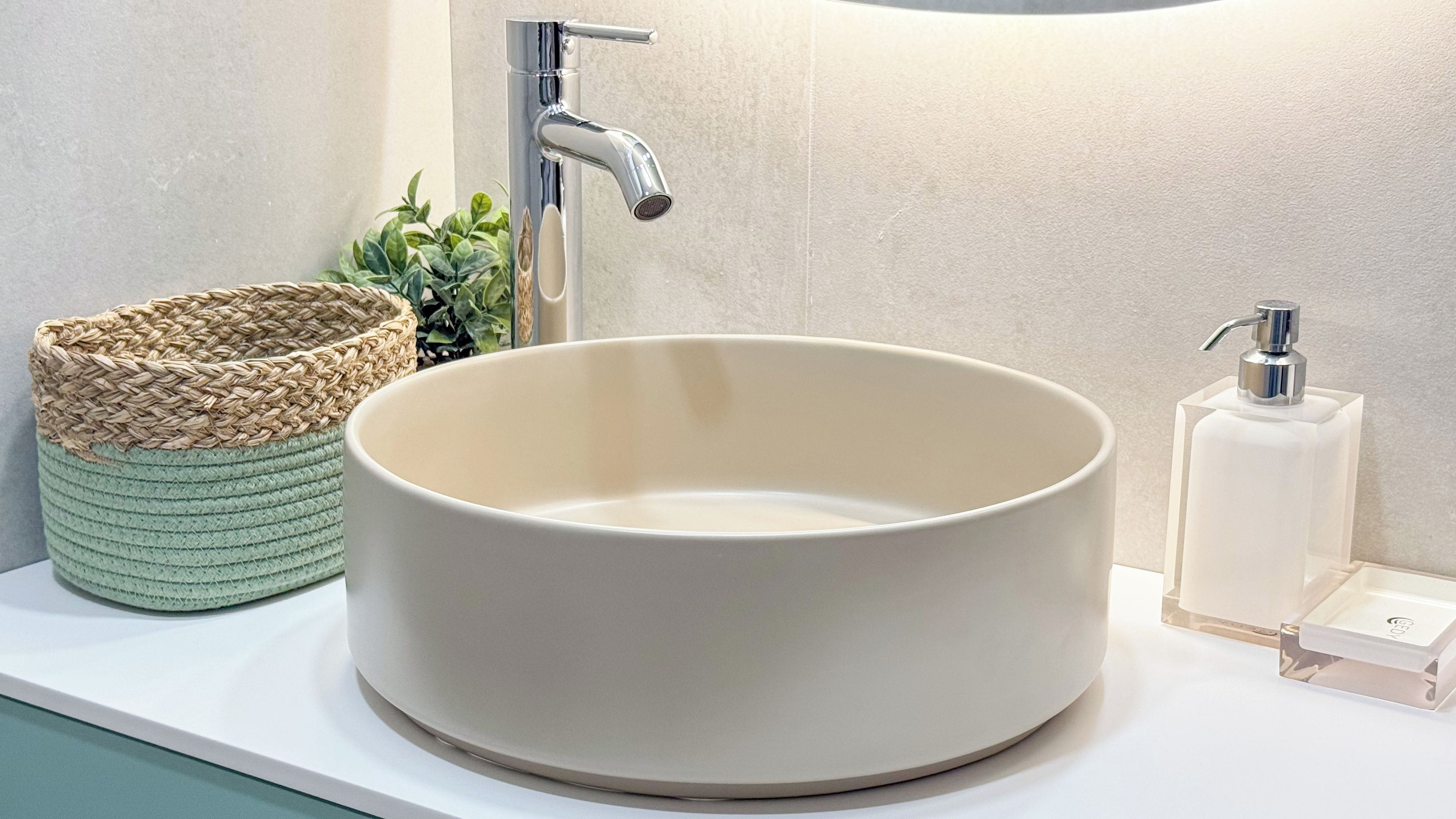 A bathroom with a round ceramic sink, a woven basket, a small plant, and a soap dispenser.