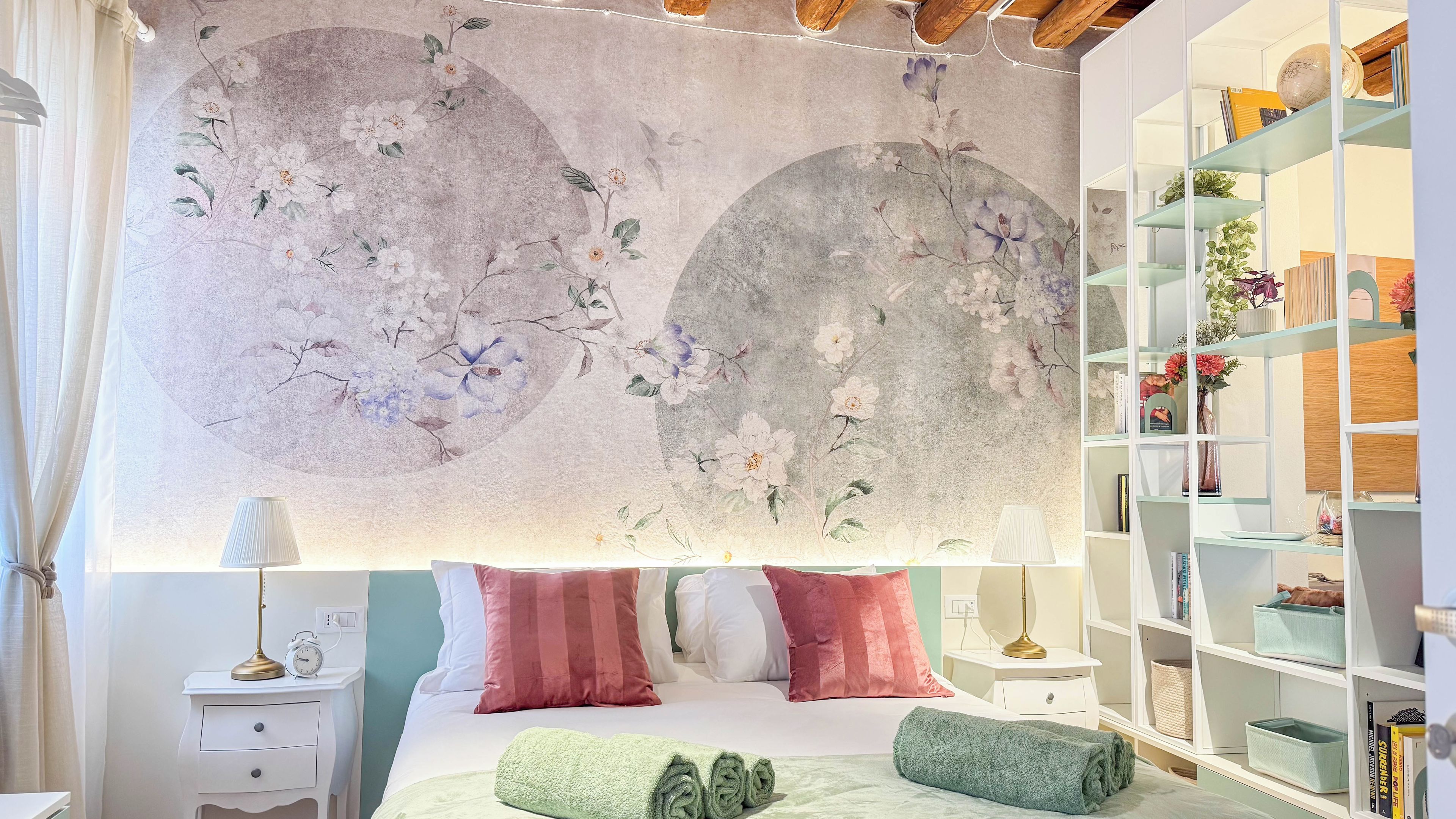 Quarto com um grande mural floral, uma cama com almofadas, uma mesa de cabeceira com abajur, um criado-mudo e uma estante de livros.
