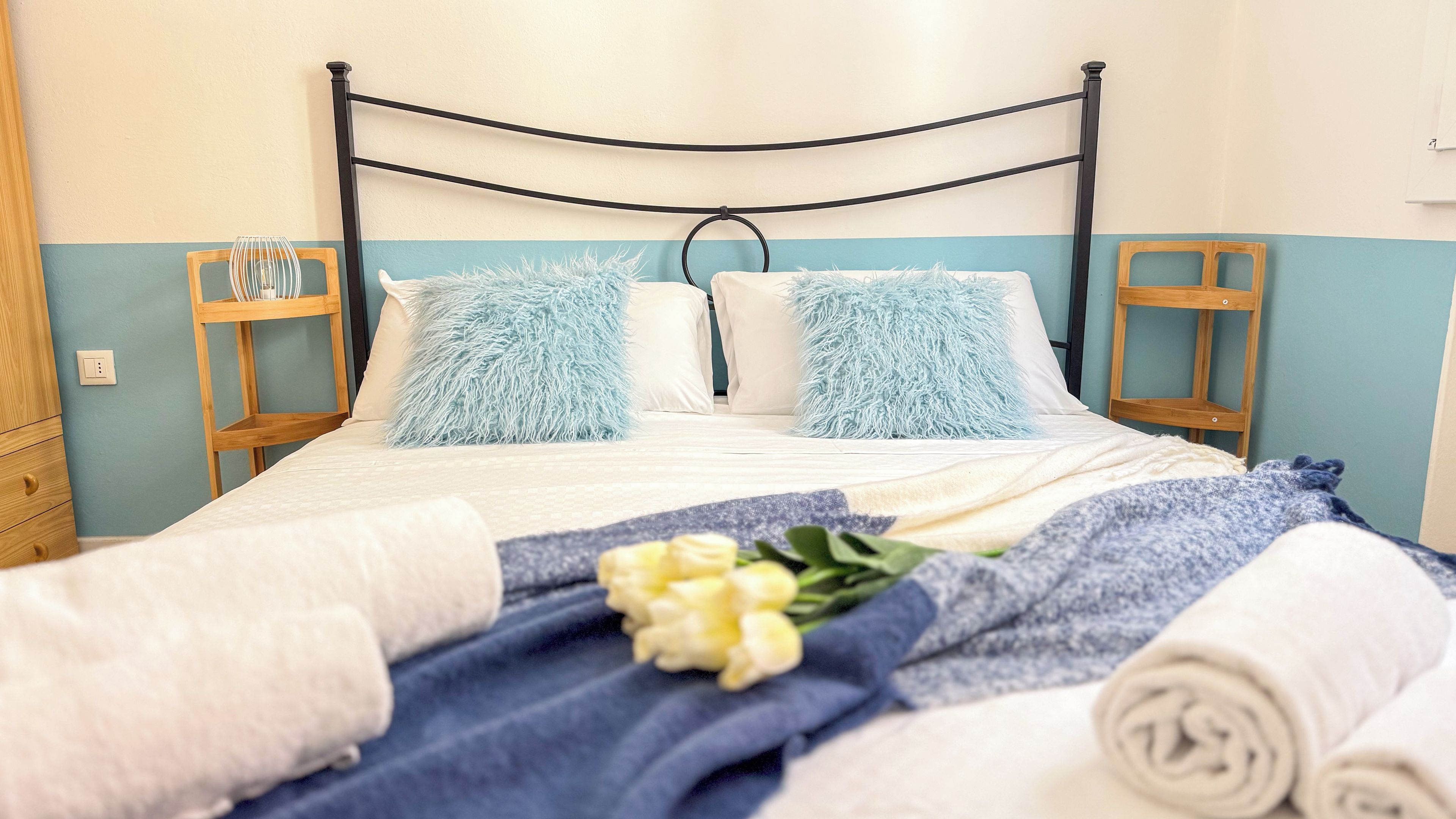 Una cama con estructura de metal negro, dos almohadas mullidas, una almohada blanca y una manta azul.