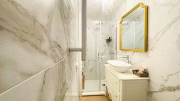 A bathroom with a marble wall, a white sink, a gold-framed mirror, and a shower enclosure.