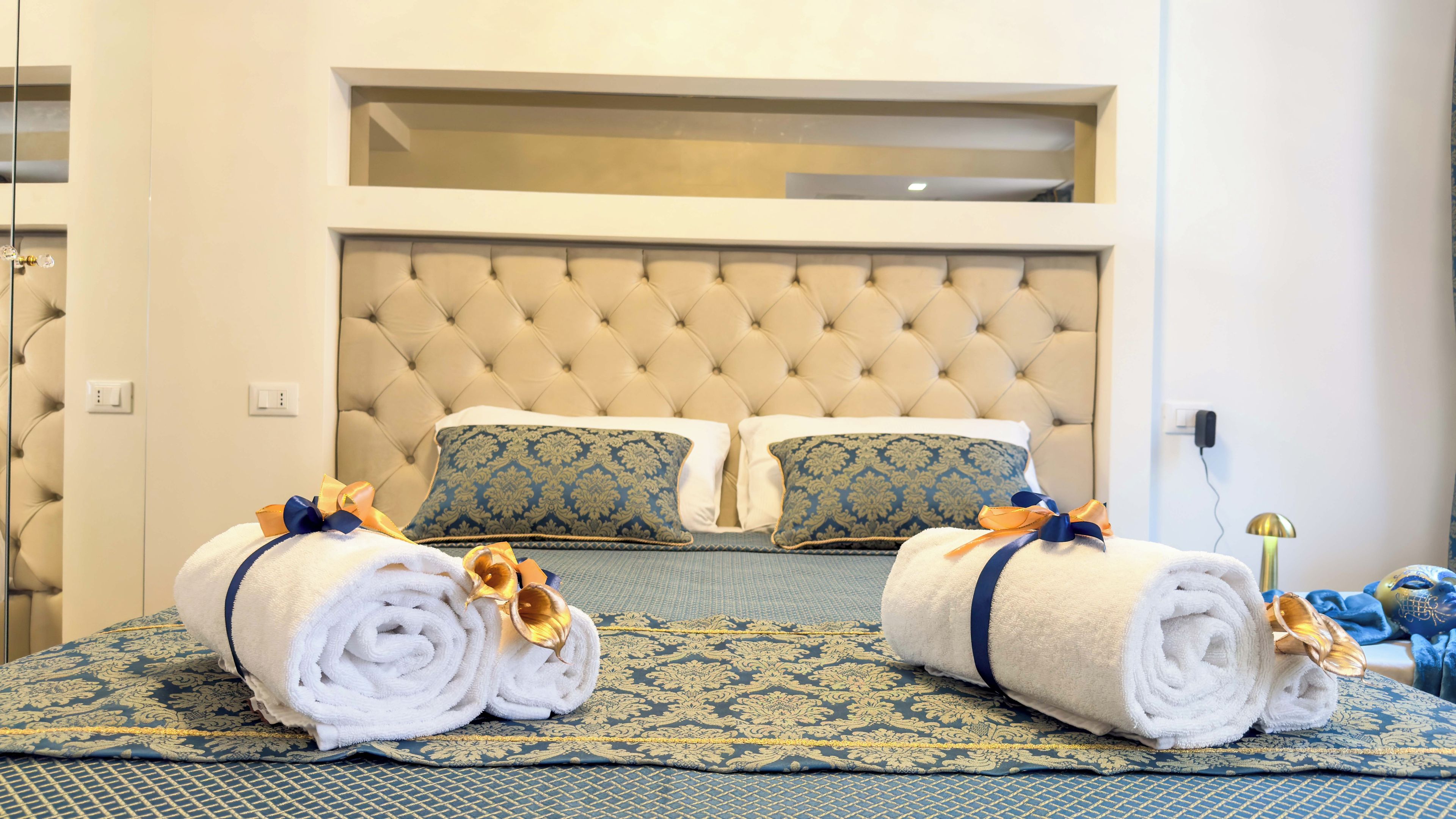 Ein Hotelzimmer mit einer gebügelten Kopfstütze, zwei Kissen mit blau-goldenem Muster, eine Bettdecke ebenfalls mit blau-goldenem Muster und zwei gerollte Handtücher mit goldenen Schleifen.
