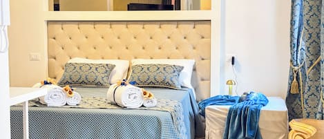 A hotel room with a tufted headboard, two beds with patterned bedding, a bedside table with towels, and a nightstand with a lamp.