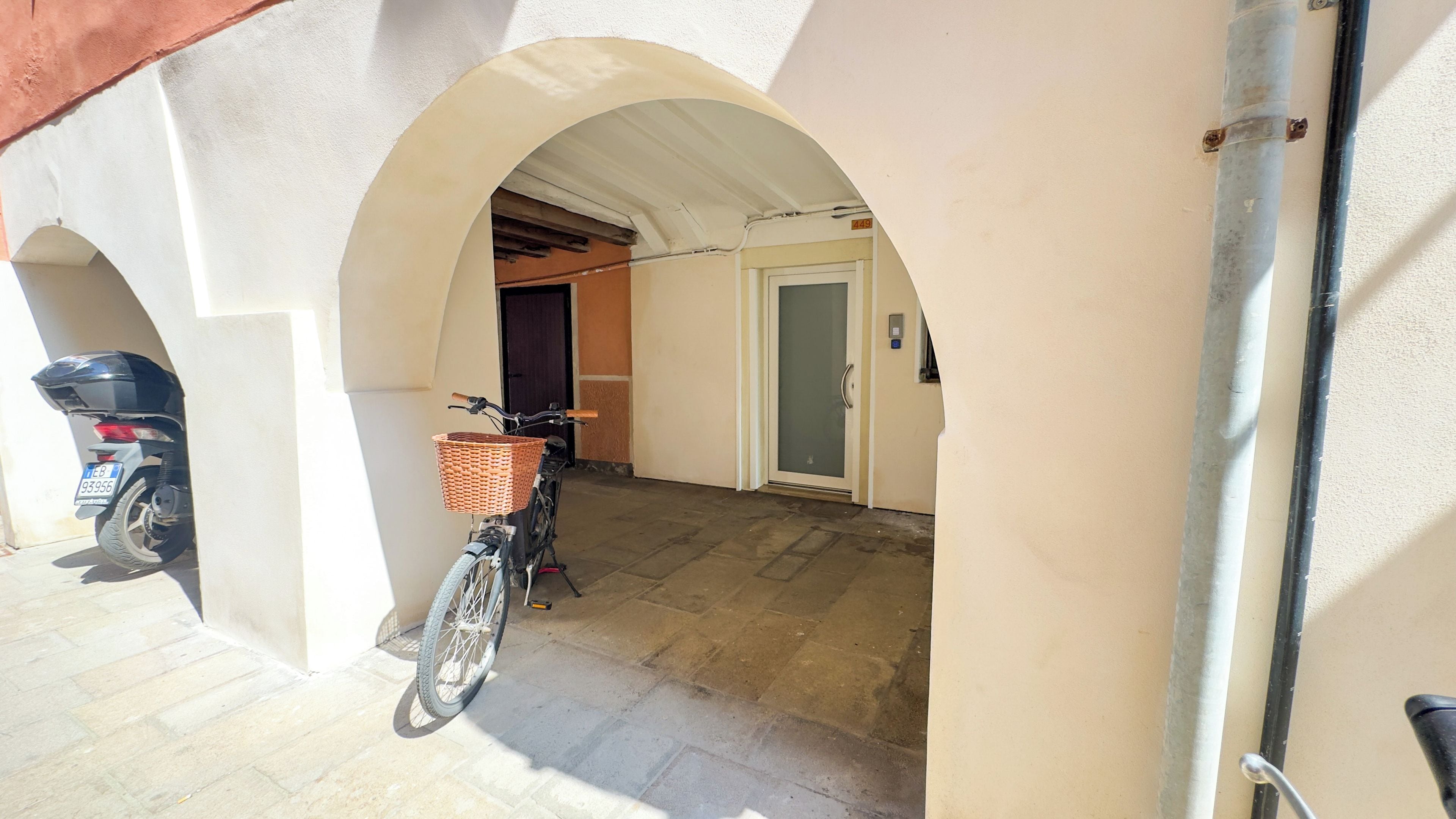 A covered outdoor area with a bicycle and a motorcycle parked under an archway, leading to a door with a number plate.