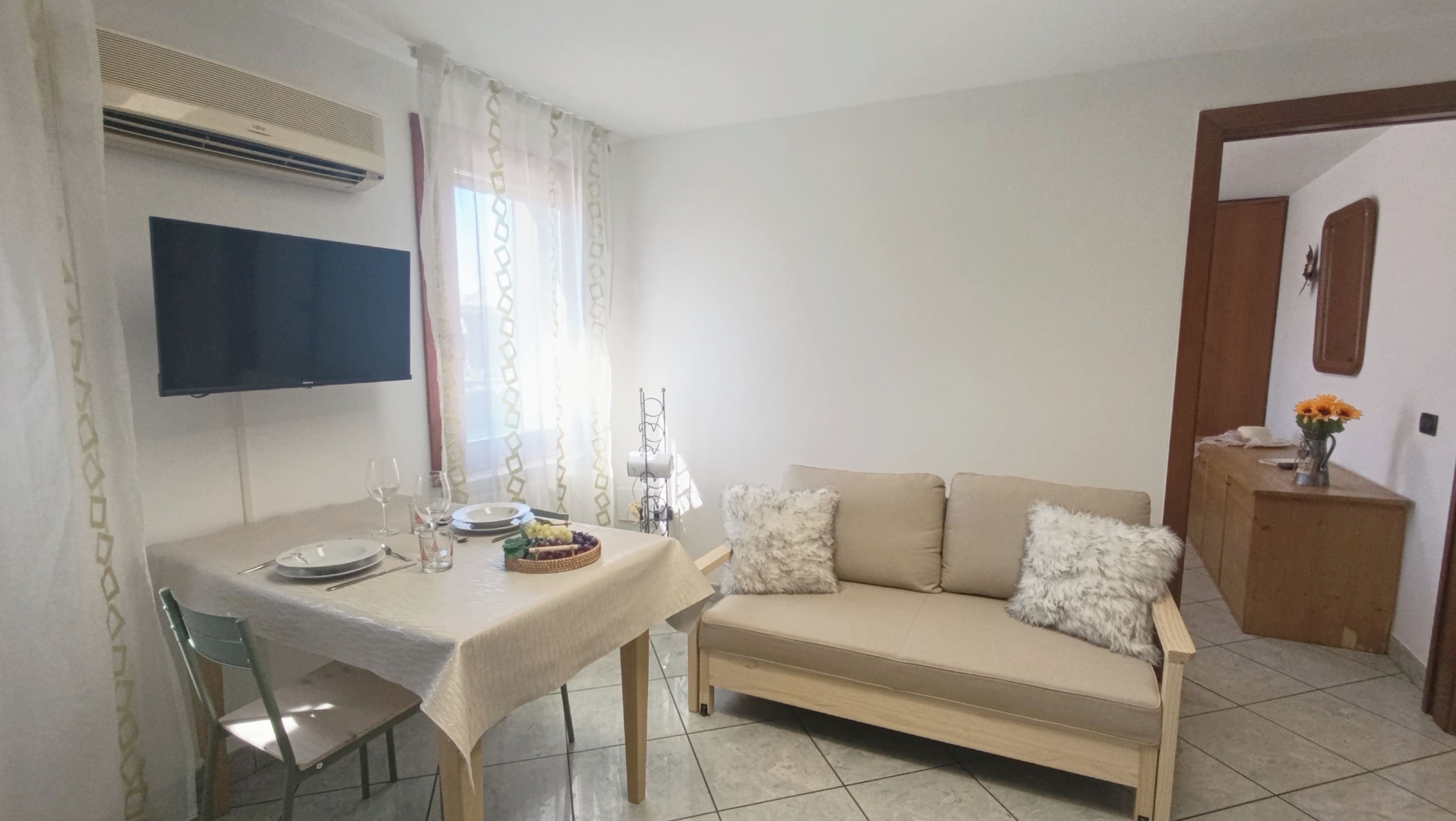 Un comedor con una mesa dispuesta para dos personas, un sofá y un mueble bajo con un jarrón de flores.