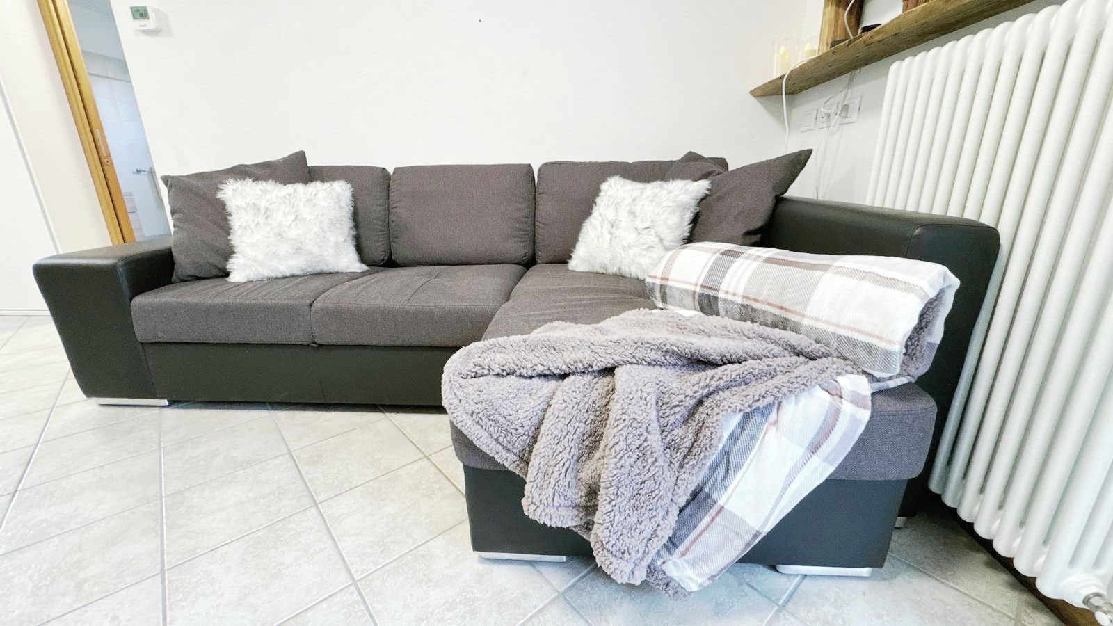 A grey sofa with two white fluffy pillows and a grey blanket on a black ottoman.