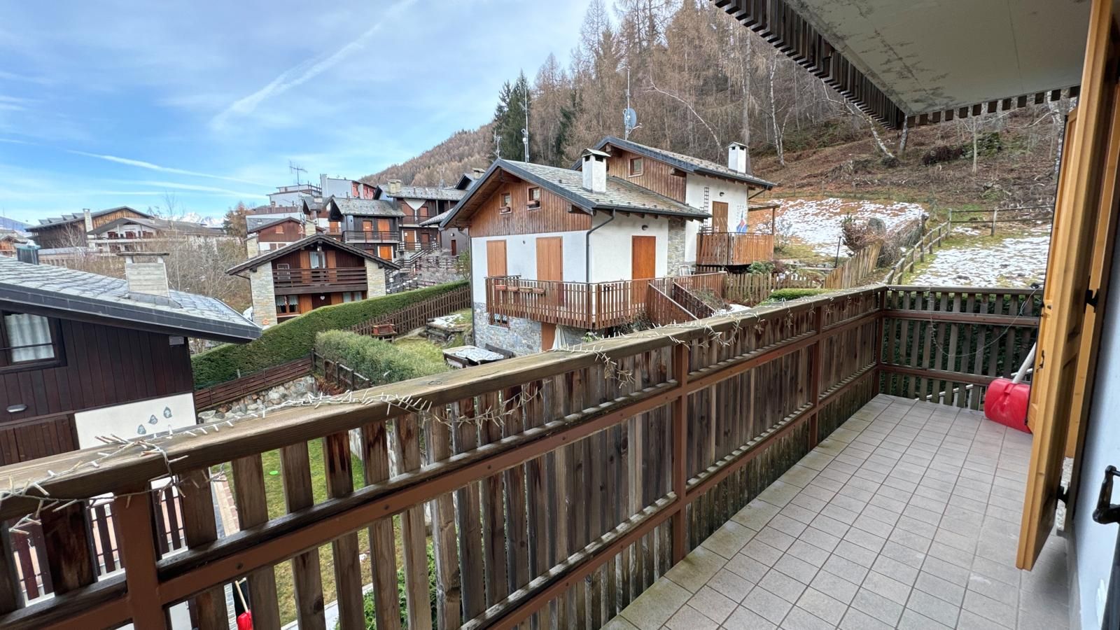 A balcony with a wooden railing overlooking a residential area with houses and a mountain backdrop.