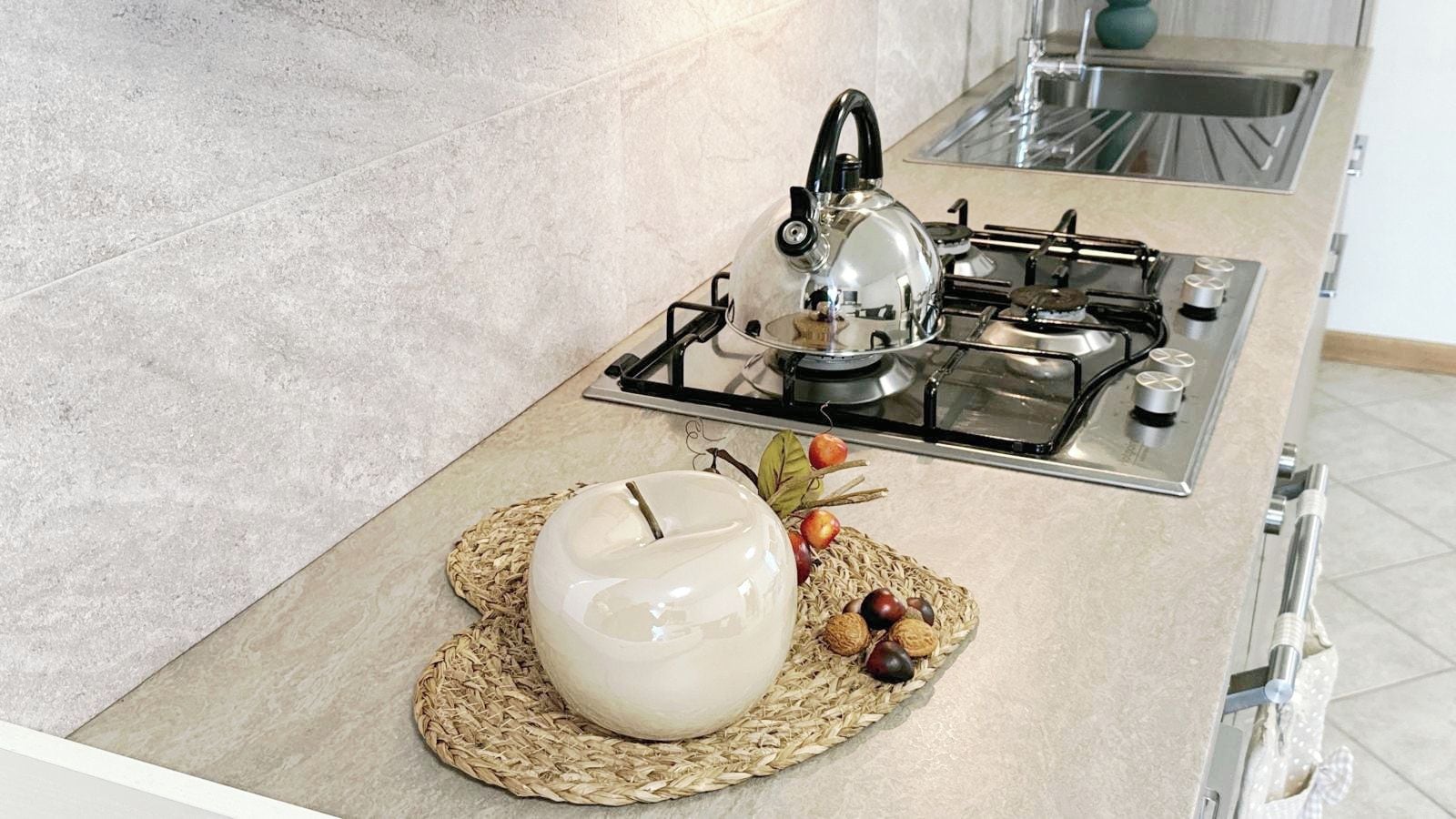 A kitchen with a gas stove, a kettle, and a decorative bowl on a woven mat.