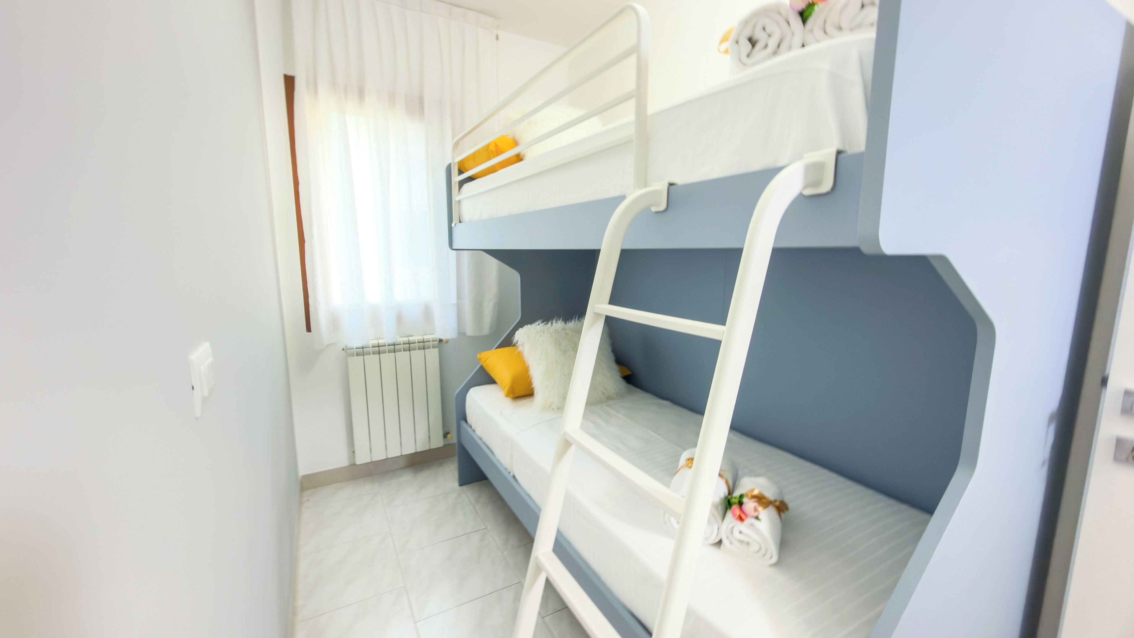 Camera con letto a castello, una scala bianca, un termosifone e una finestra con tende.