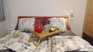 Uma cama com um cobertor branco e fofo, um tabuleiro com uma chaleira vermelha, duas canecas e um prato com comida.