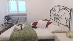 Ein ordentlich bezogenes Bett mit einer grünen Decke, einem weißen Kissen und einem roten Kissen. Es gibt ein Fenster mit Vorhängen, eine Heizung und einen Stuhl mit Blumenmuster.