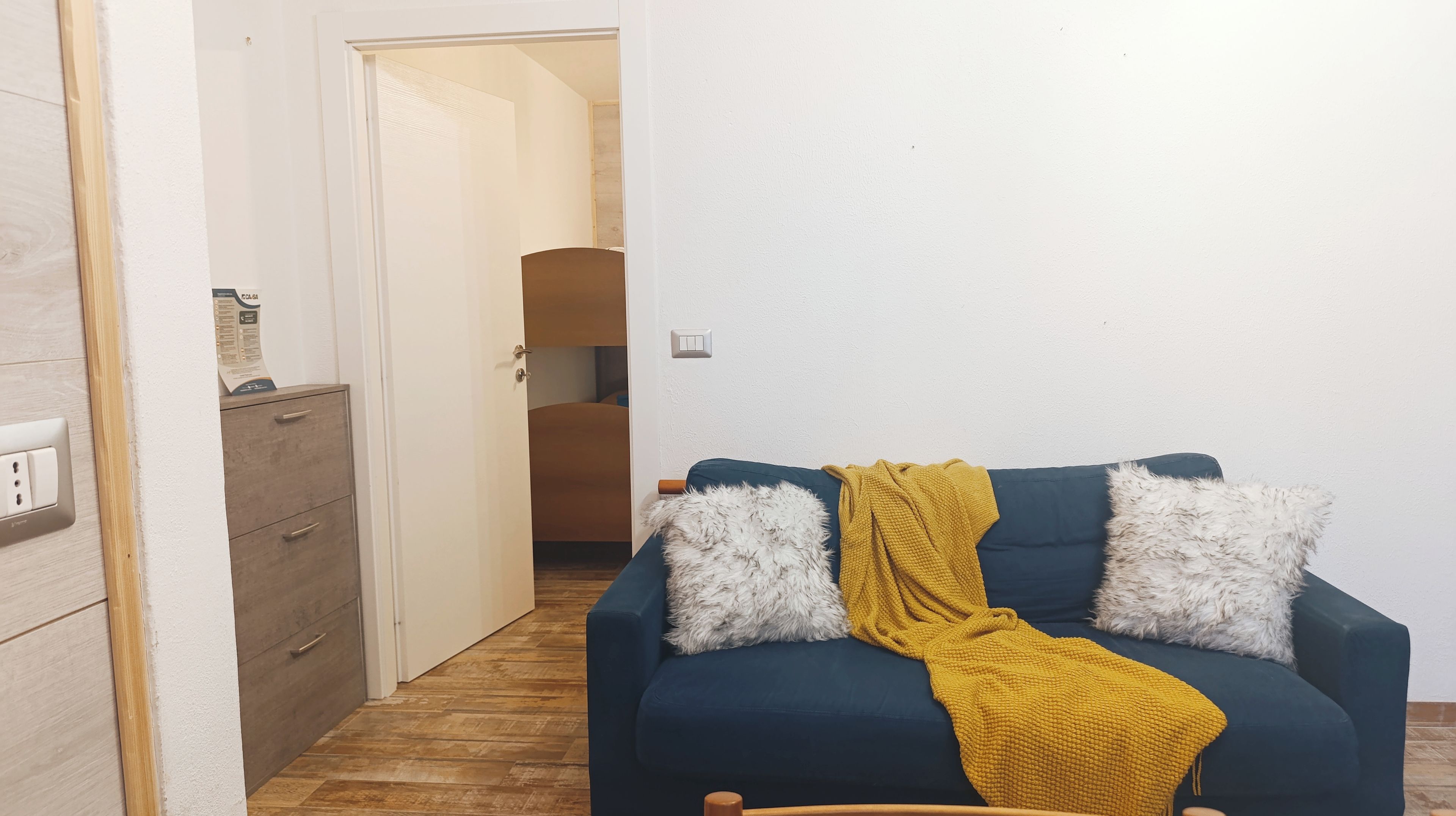 A room with a blue sofa, two white fluffy pillows, a mustard-colored throw blanket, and a wooden wardrobe.
