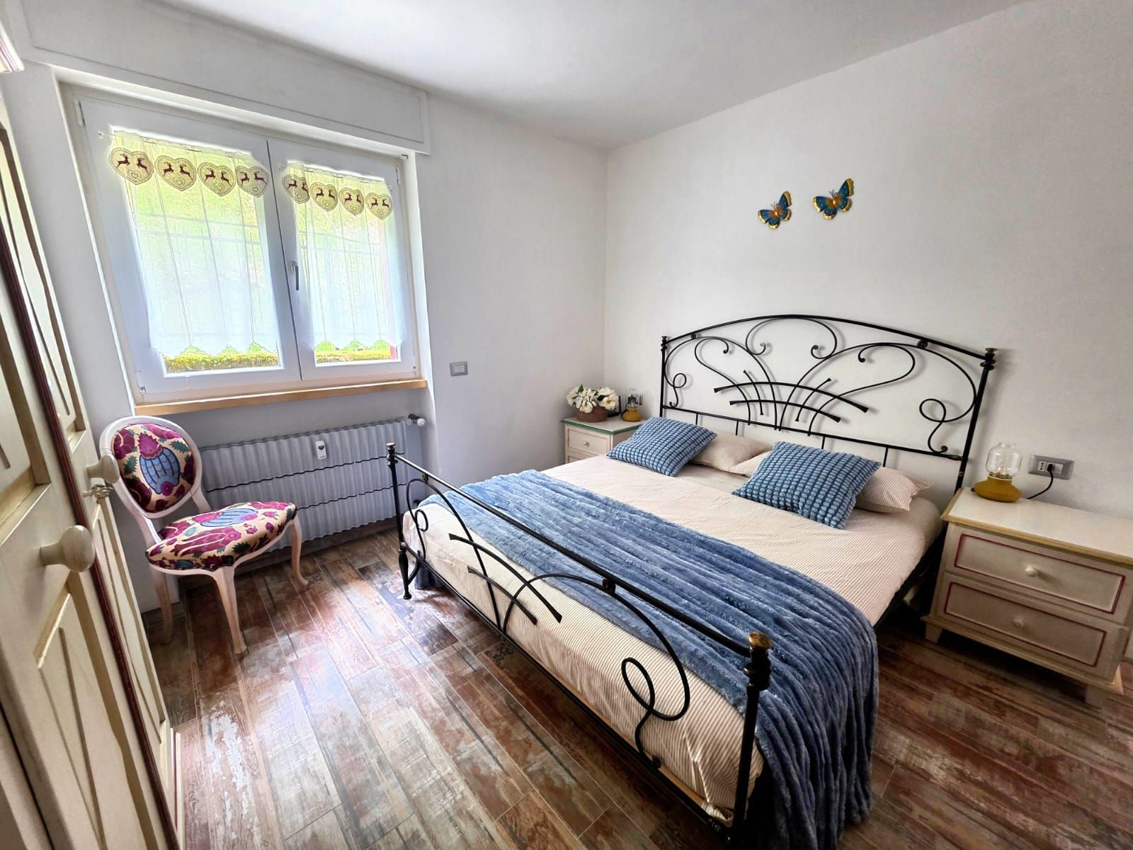 A bedroom with a bed, a chair, a window with curtains, a bedside table, and a wall decoration.