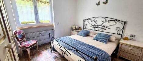 Un dormitorio con cama, una silla, una ventana con cortinas, una mesita de noche y un adorno en la pared.
