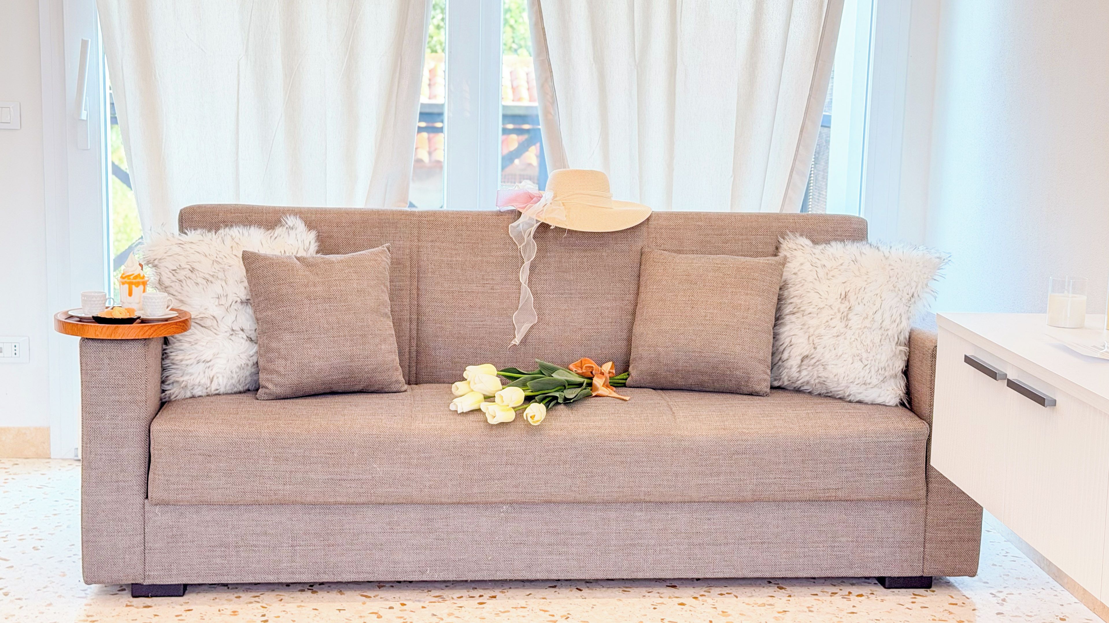 A beige sofa with fluffy pillows, a tray with a teapot and cups, and a bouquet of flowers.