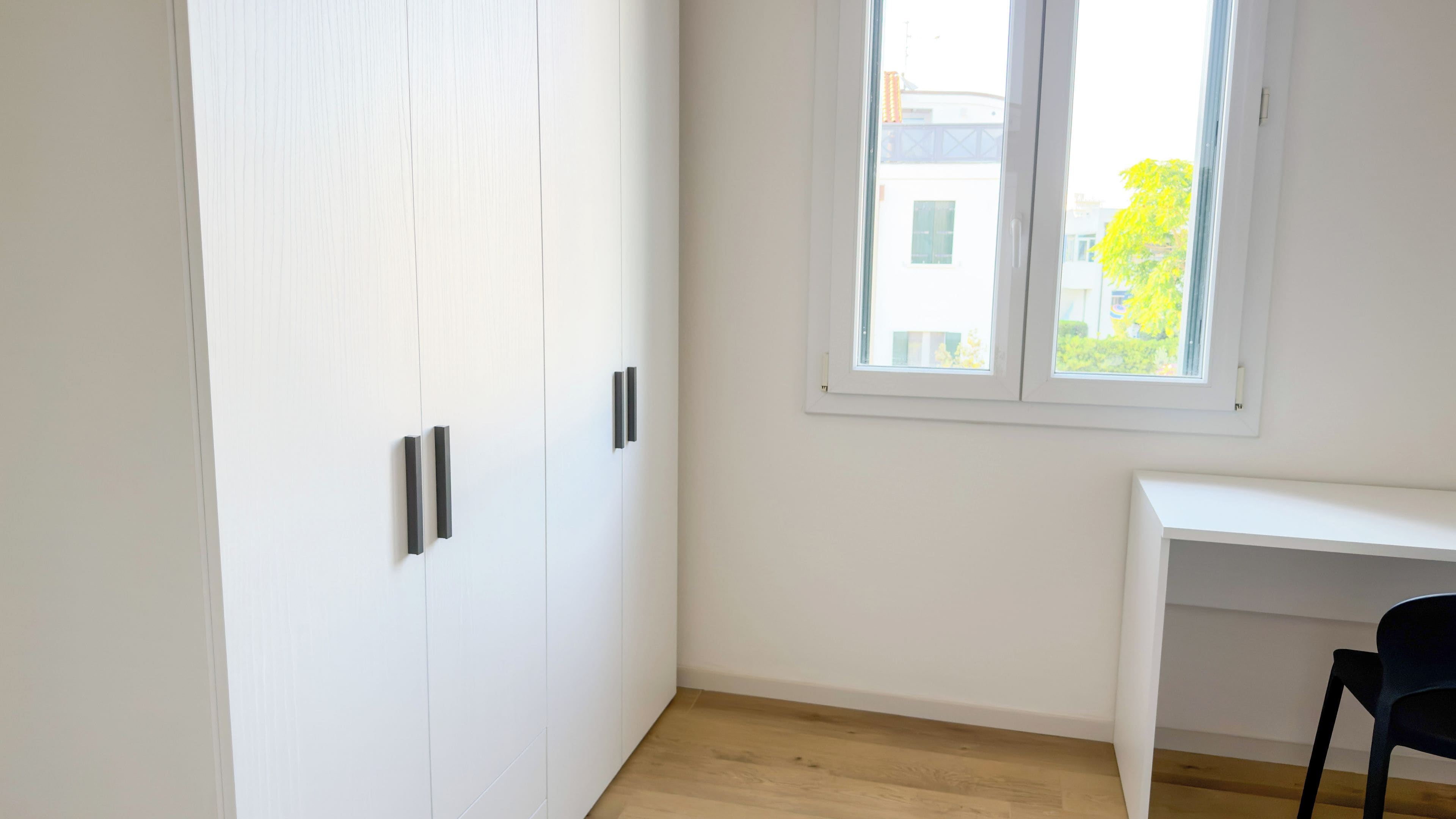 A room with a white wardrobe, a white desk, and a window with a view of buildings and trees.