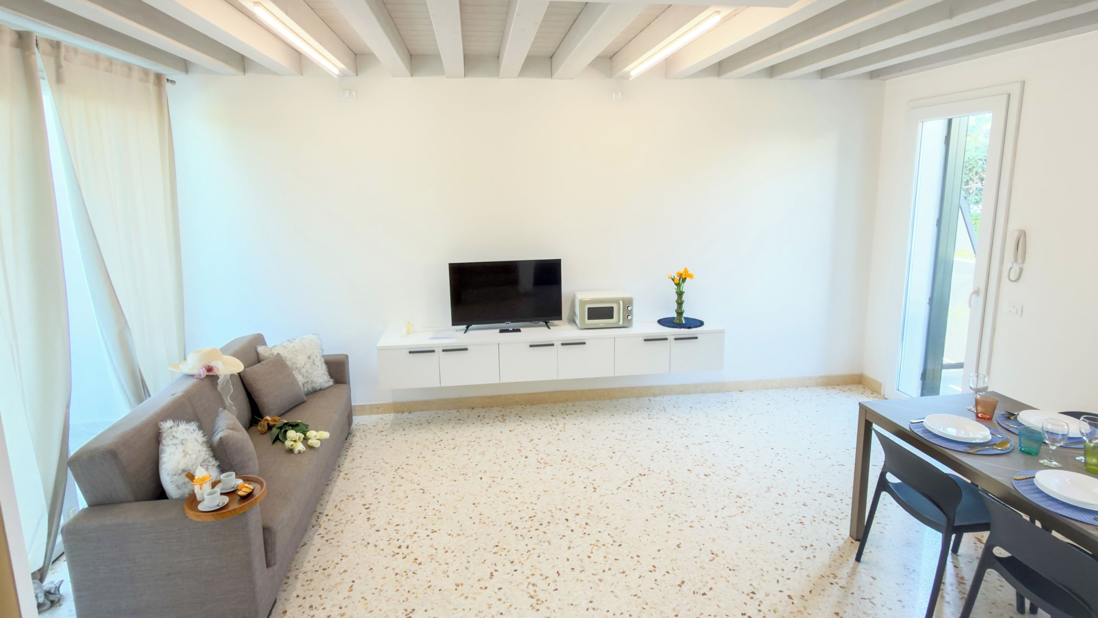 A modern living room with a grey sofa, a flat-screen TV, a microwave, and a dining area with a table set for two.