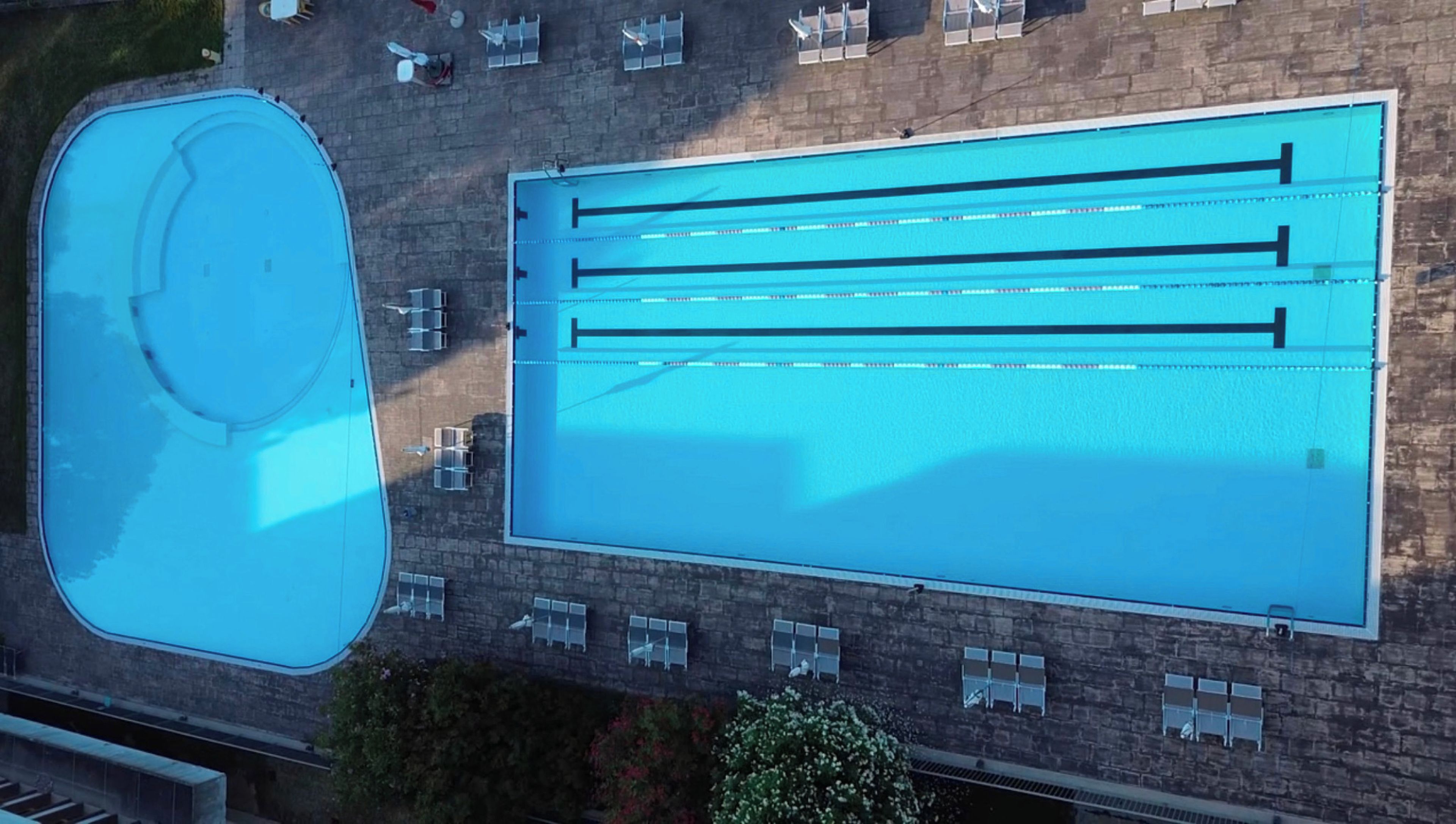 Vista aérea de una piscina con múltiples carriles, una piscina más pequeña separada y un patio de ladrillo con mobiliario exterior.