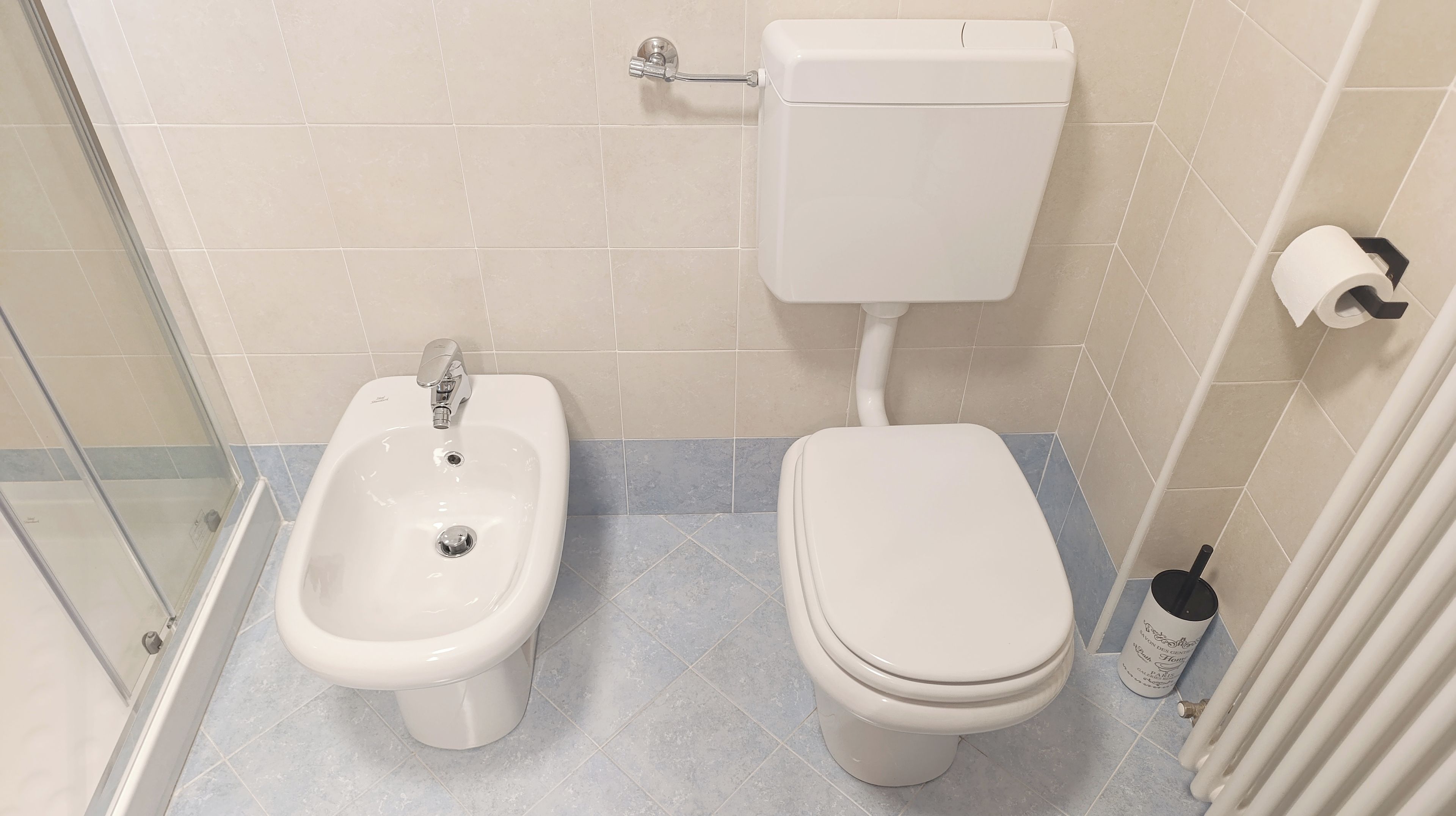 A bathroom with a bidet, toilet, and a roll of toilet paper on the wall.