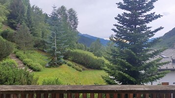 Paisagem montanhosa com um deck de madeira que dá para uma área gramada com árvores e um edifício com telhado de azulejos.