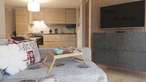 Un salon moderne comprenant un canapé, une table basse sur laquelle est posé un plateau de petits gâteaux, un coin cuisine avec des meubles en bois et un téléviseur à écran plat fixé au mur.