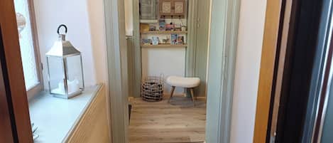 A narrow hallway with a wooden floor, a wicker basket, a small stool, and a shelf with books and decorative items.