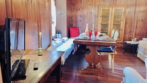 Ein Holzinnenraum mit einem für vier Personen gedeckten Esstisch, einem weißen Sofa, einem roten Kissen und einem Fernseher auf einem Holzboden.