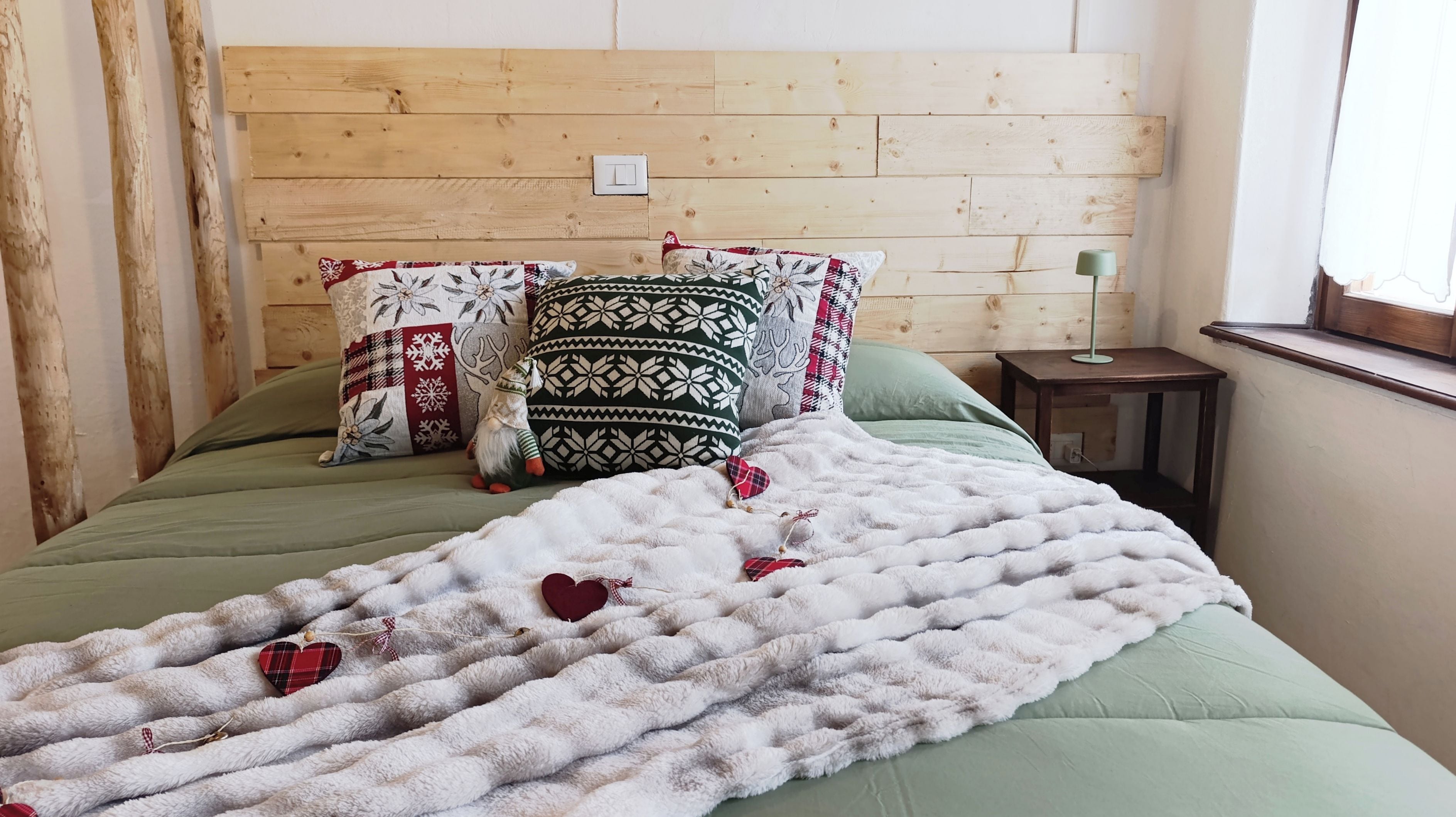 A bed with a green bedspread, a white fluffy blanket with red hearts, and patterned pillows.