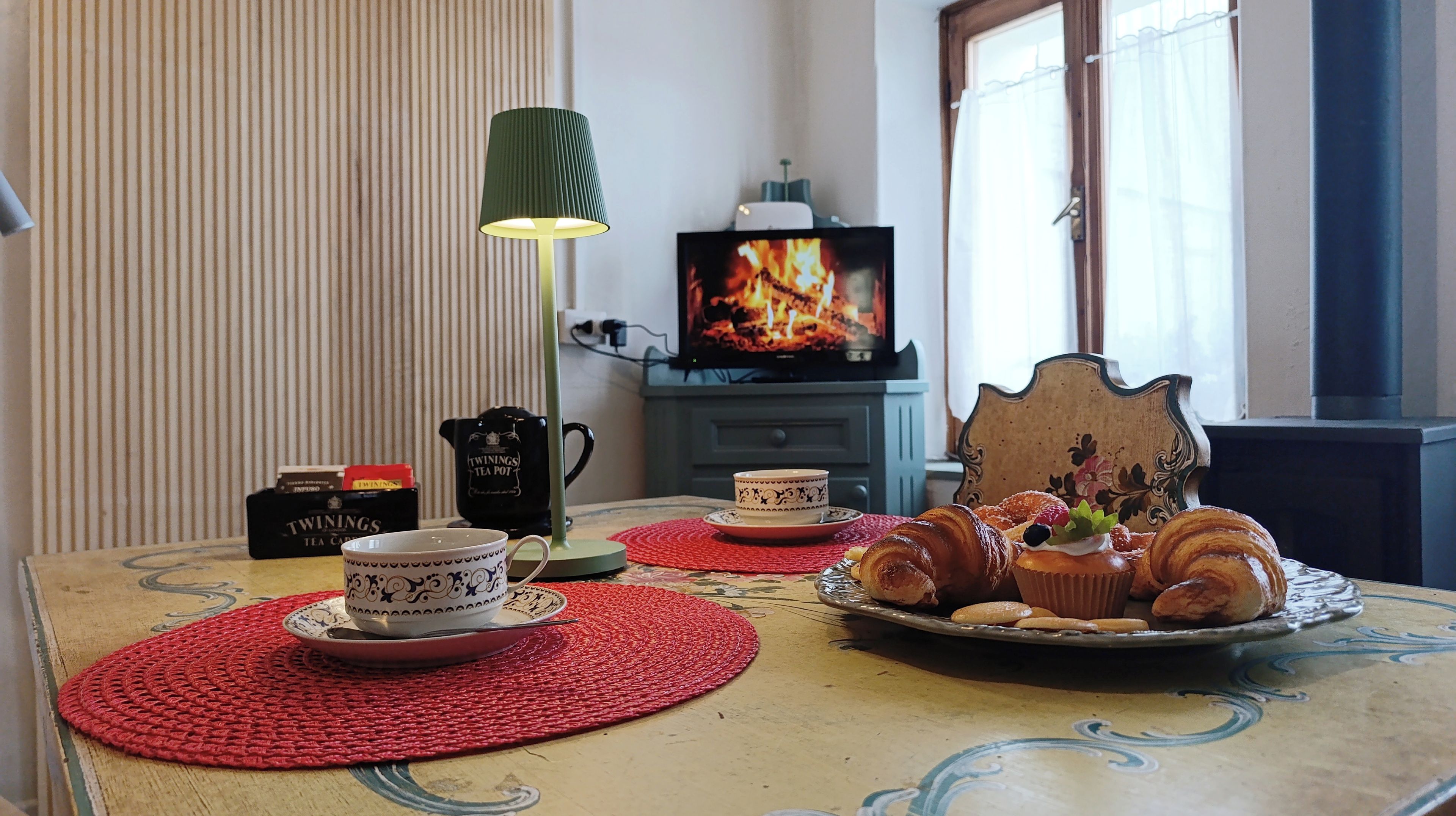 Un salon chaleureux avec un téléviseur diffusant une image de feu de cheminée, une table basse sur laquelle sont disposées des pâtisseries et des tasses à thé, et un fauteuil décoratif.
