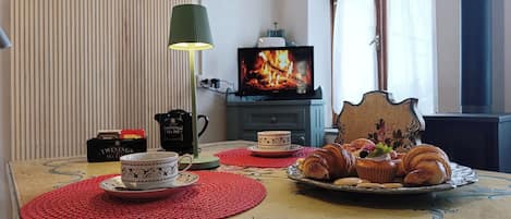 Una sala de estar acogedora con un televisor mostrando un fuego, una mesa de centro con pasteles y tazas de té, y un sillón decorativo.