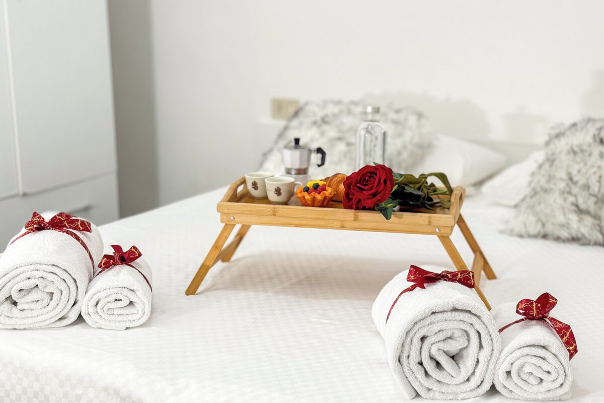 Ein ordentlich gemachteres Bett mit aufgerollten Handtüchern, einem Tablett mit Teekanne, Tassen, einer Rose und Gebäck.