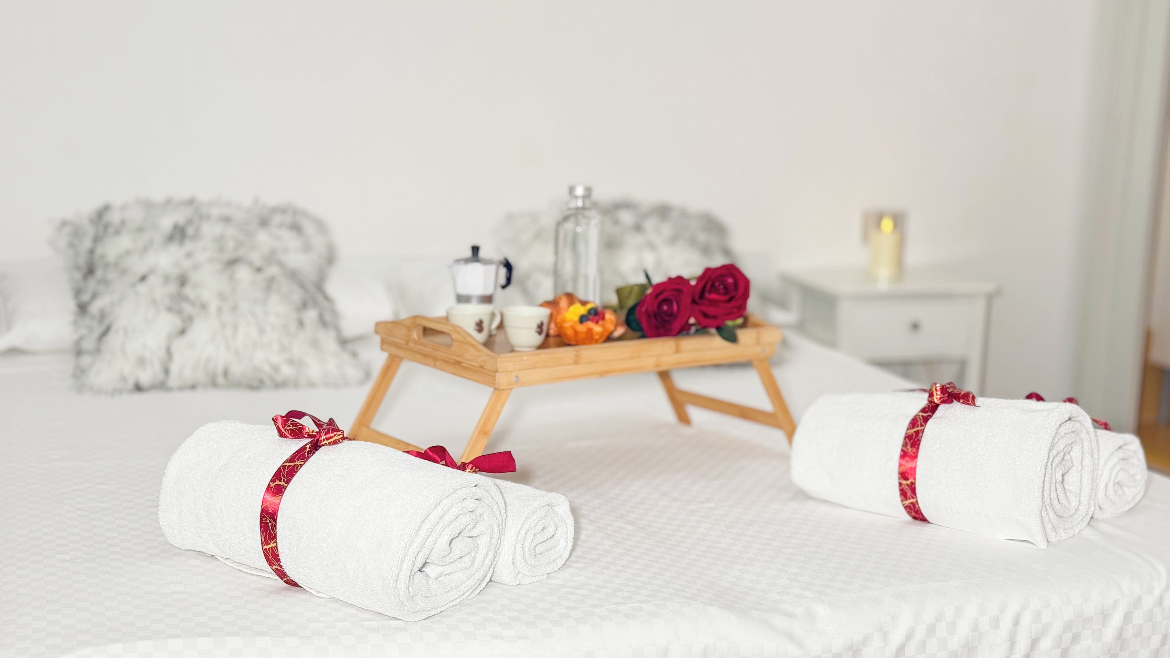 Una cama bien hecha con dos toallas enrolladas sujetas con lazos rojos, una bandeja de madera con una cafetera, tazas, un cuenco de fruta y una rosa roja.