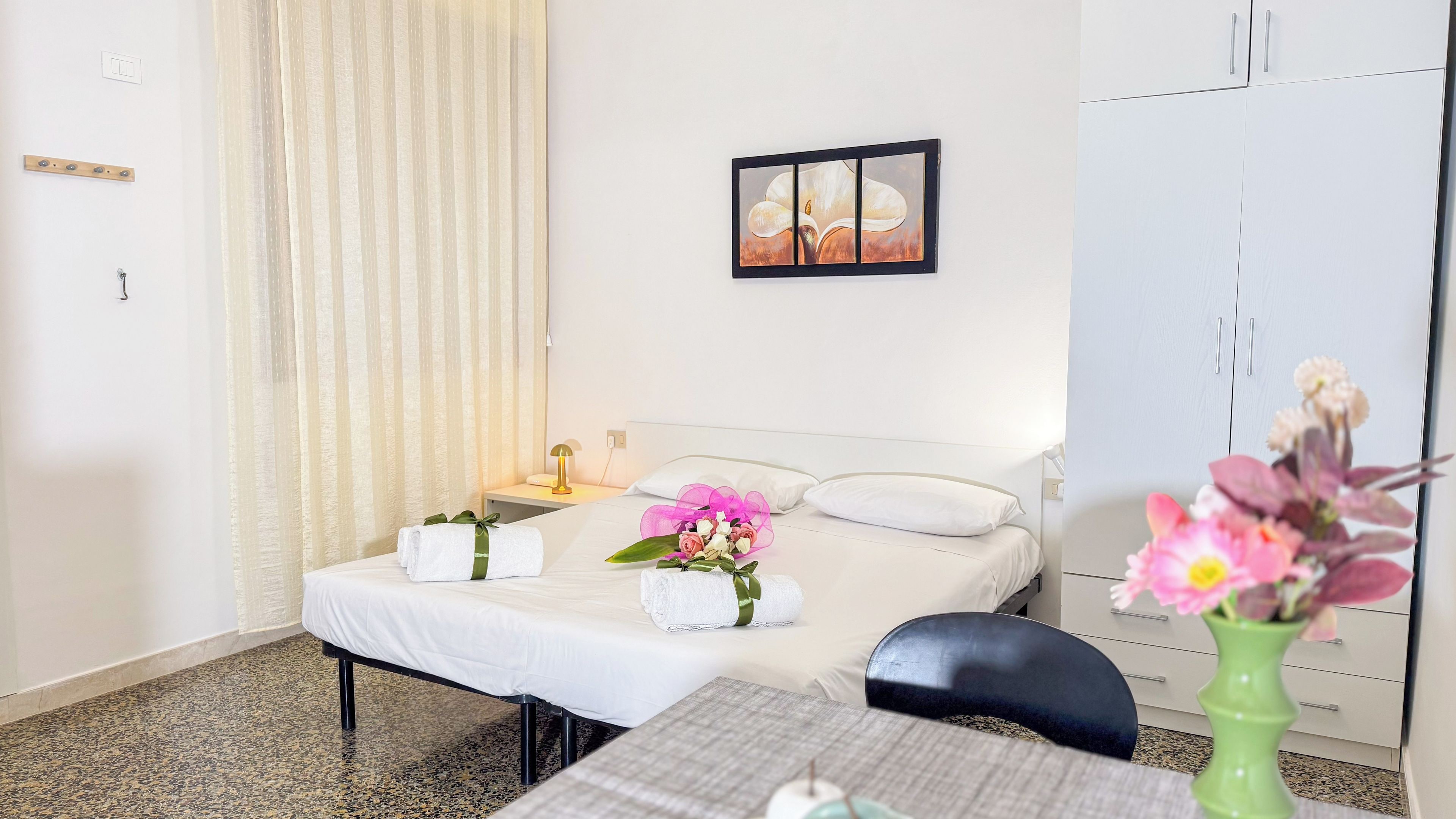 Habitación de hotel con cama, mesita de noche con lámpara, cuadro en la pared, armario y un jarrón con flores.